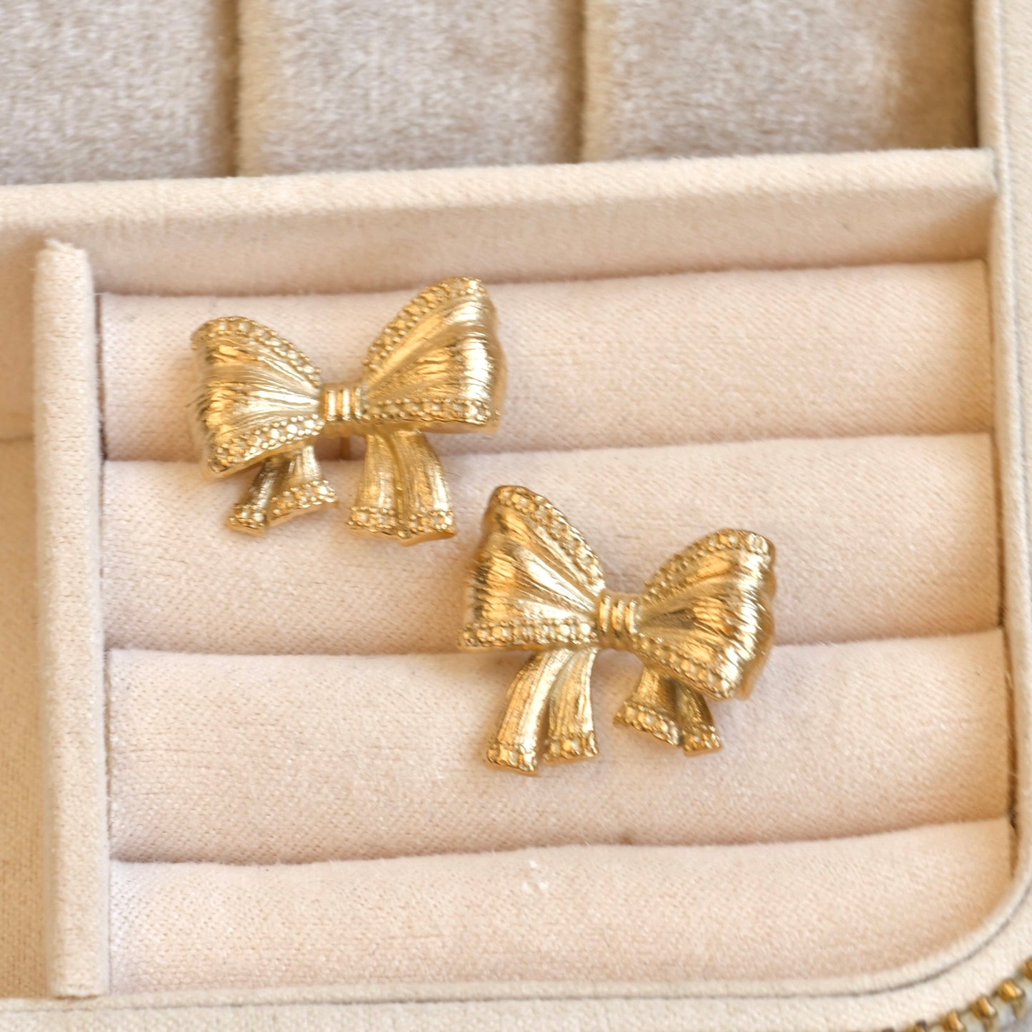Gold bow-shaped earrings on a beige jewelry display box.