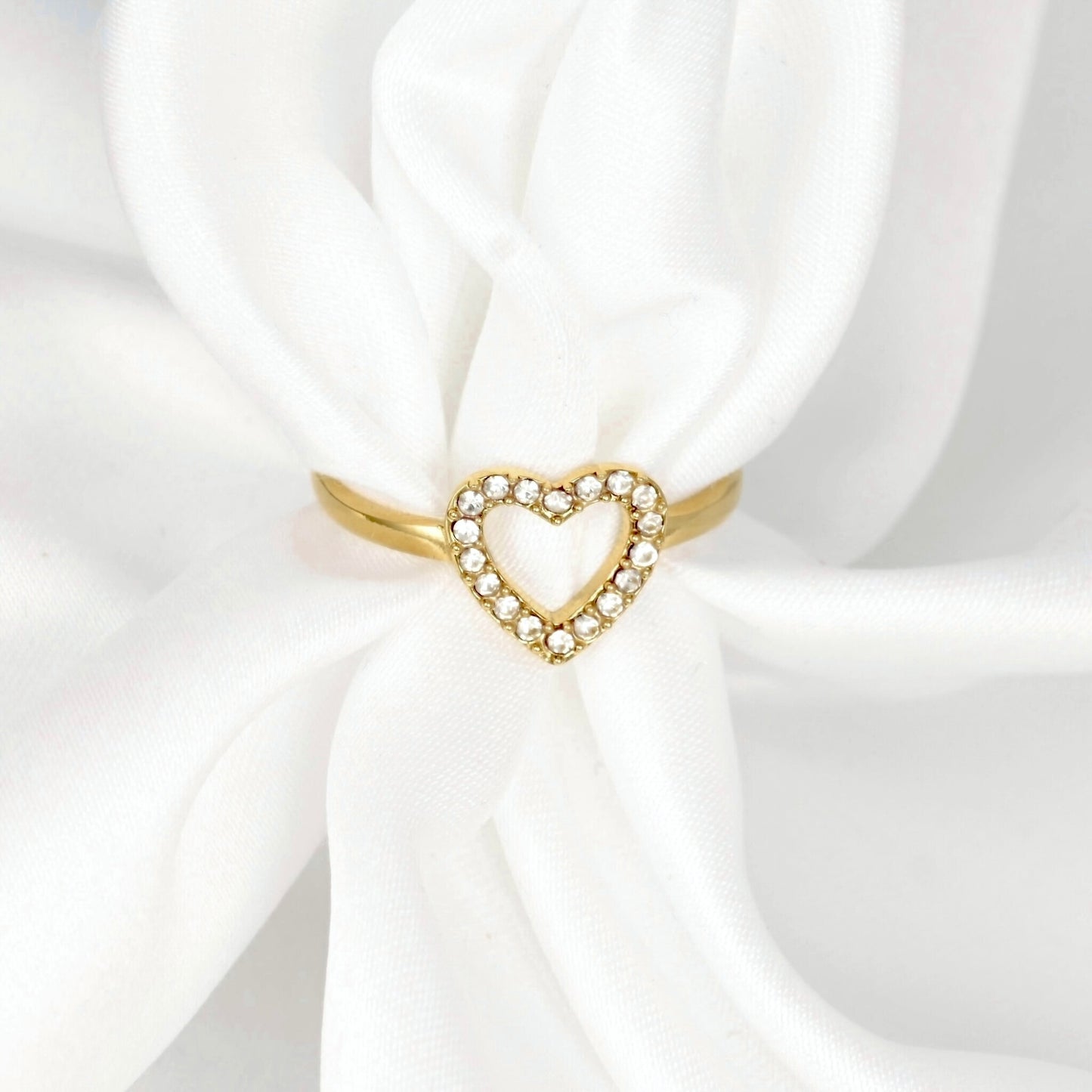A gold heart-shaped ring with a border of small clear stones, displayed on a white satin ribbon.