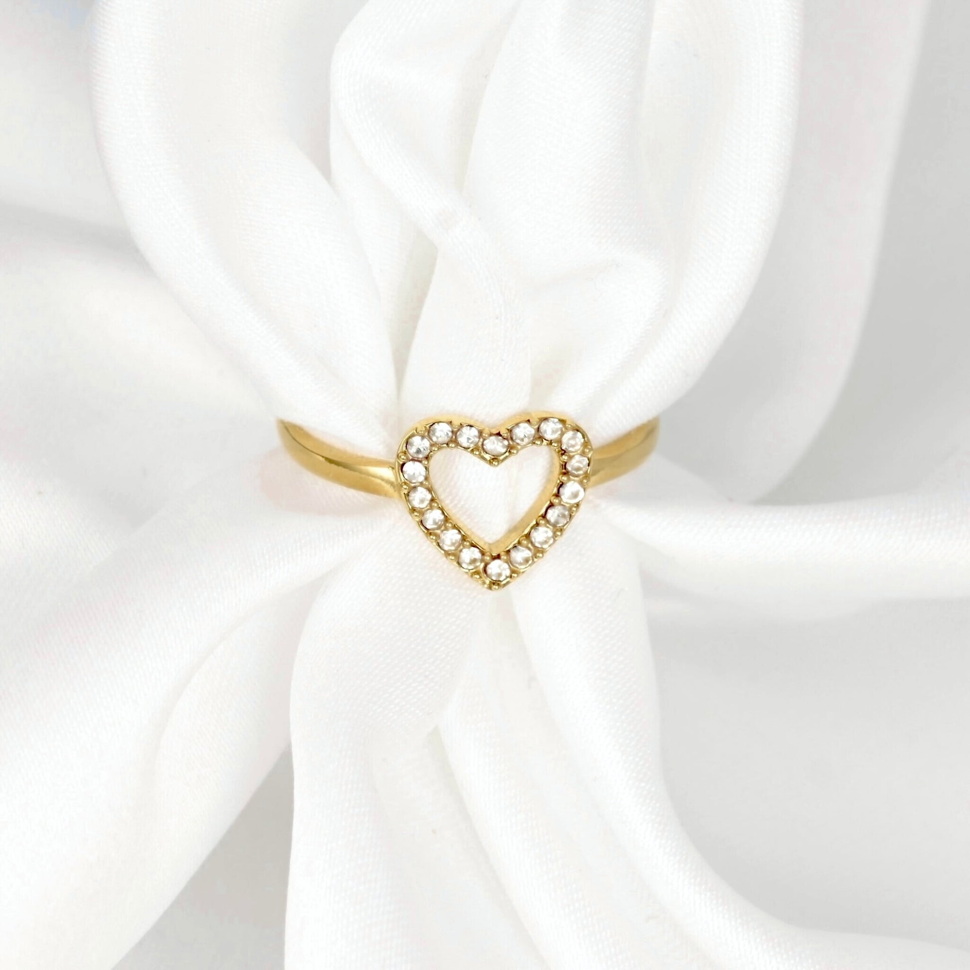A gold heart-shaped ring with a border of small clear stones, displayed on a white satin ribbon.