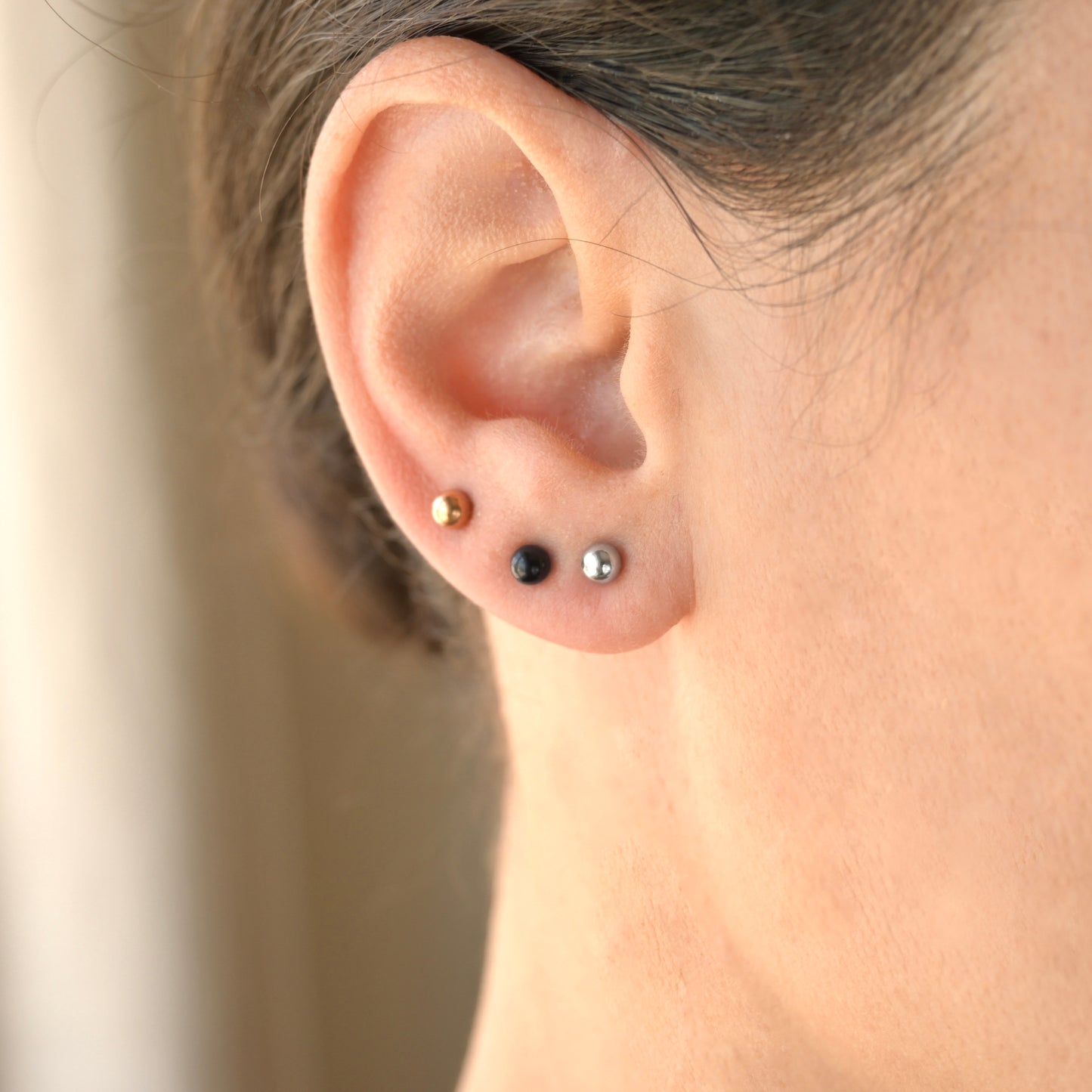 Close-up of an ear with gold, silver, and black round stud earrings against a neutral background