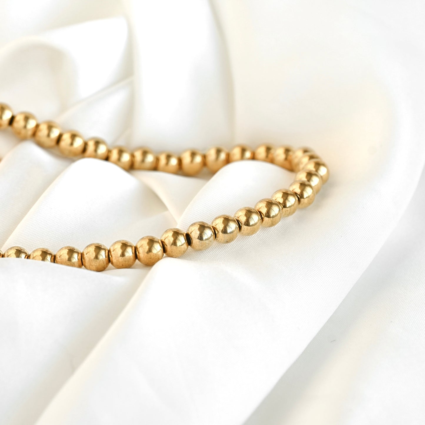 A strand of small, polished gold-colored beads draped elegantly over soft, white satin fabric.