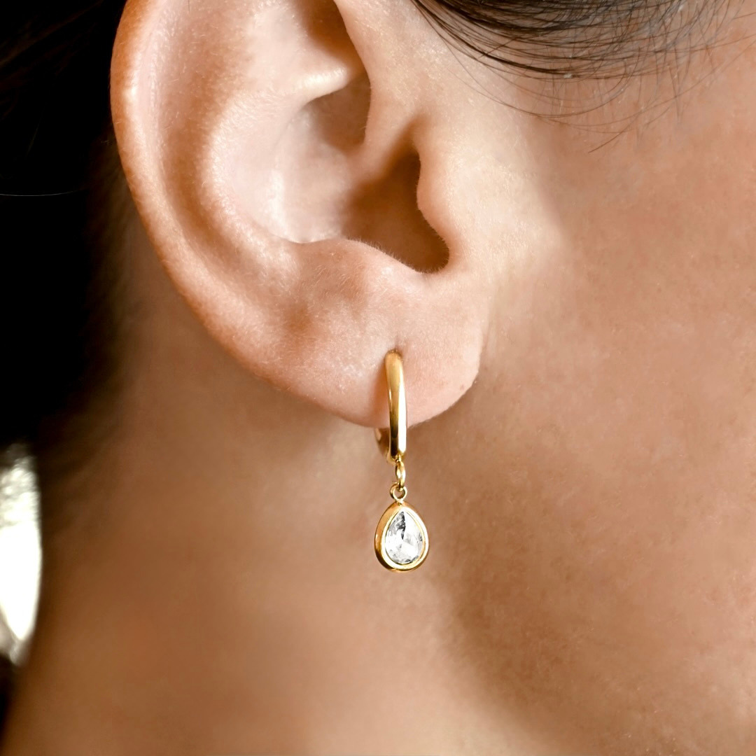 A close-up of a person's ear wearing a small gold huggie hoop earring with a teardrop-shaped clear gemstone dangle.