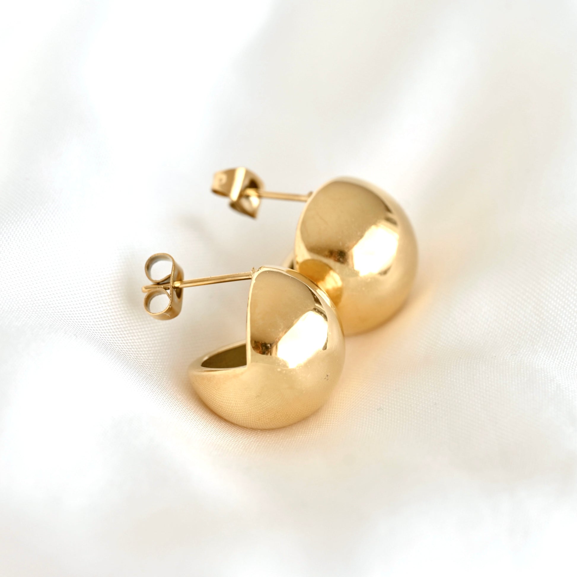 A pair of gold-colored, semi-circular dome stud earrings with post-back closures, displayed on a soft, white textured fabric background.