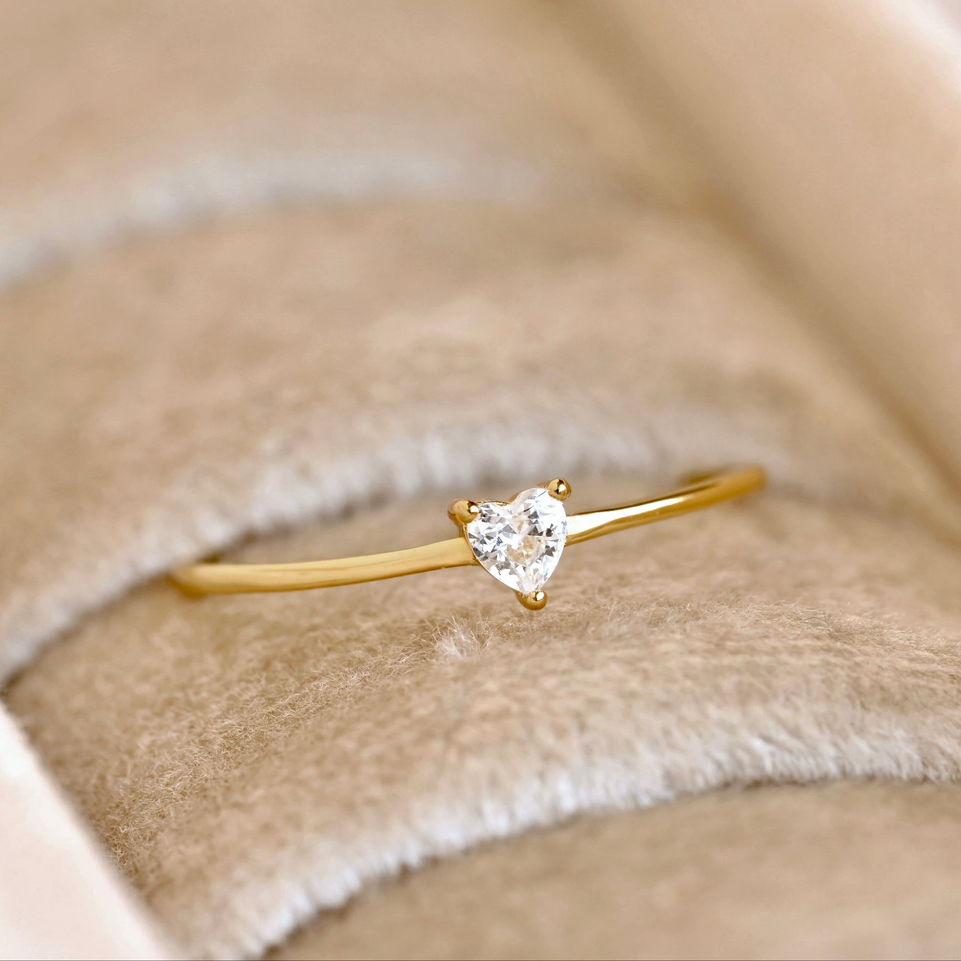 A delicate yellow gold ring with a small heart-shaped clear gemstone, displayed on a beige velvet ring box.
