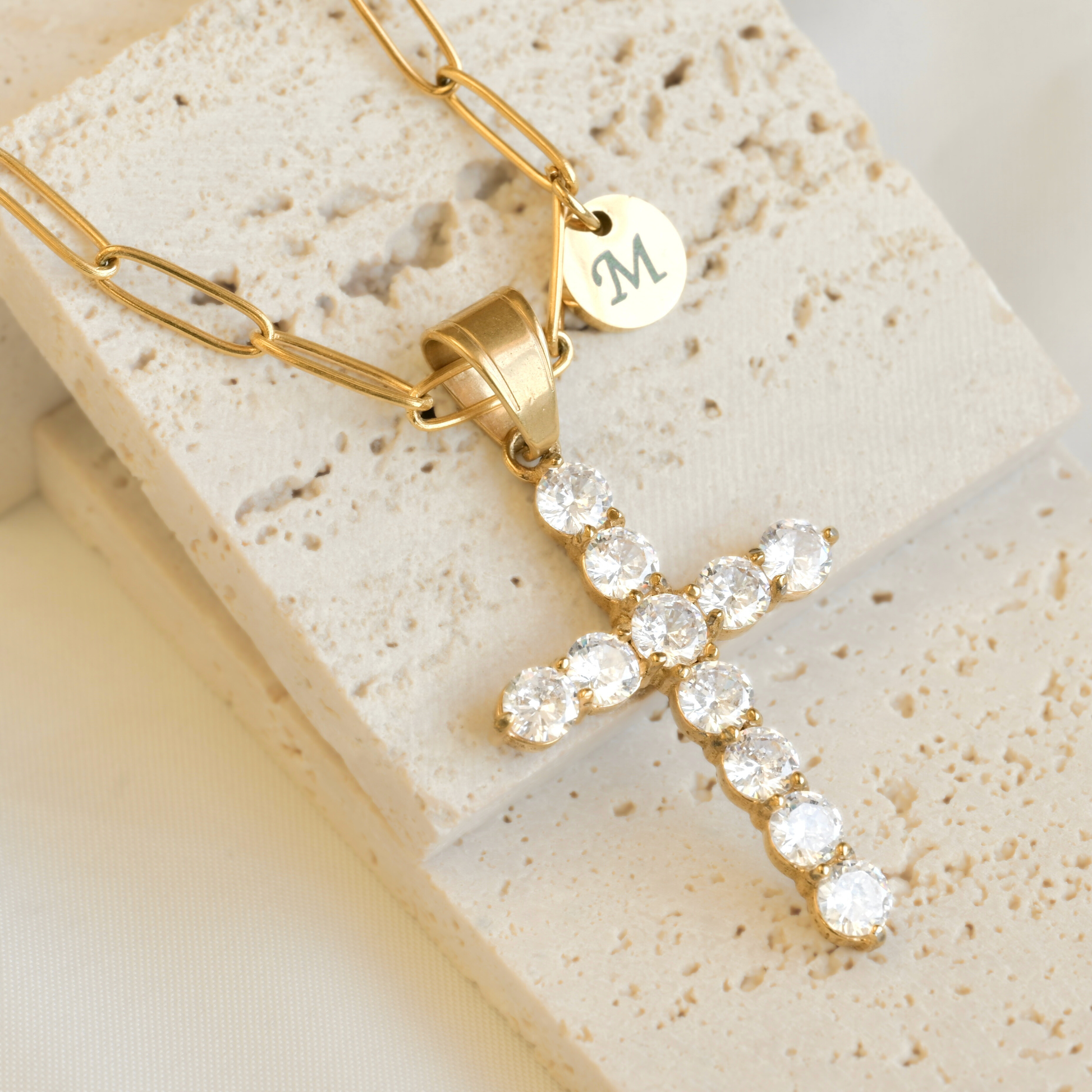 Gold  paperclip necklace with a cross pendant and a small round letter charm on a textured stone surface
