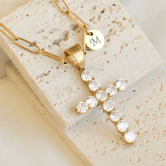 Gold  paperclip necklace with a cross pendant and a small round letter charm on a textured stone surface