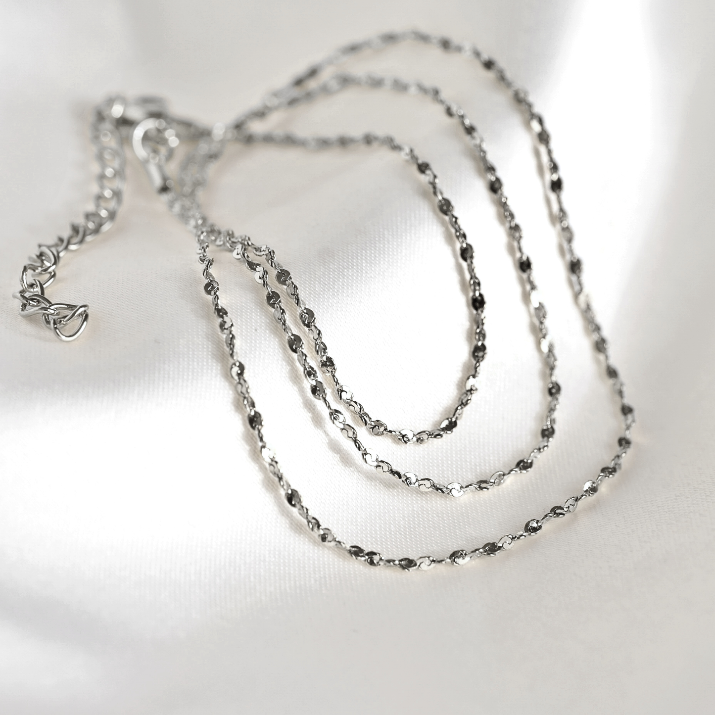 A silver-toned chain necklace with a shimmering rope link design, coiled on a white satin fabric background.