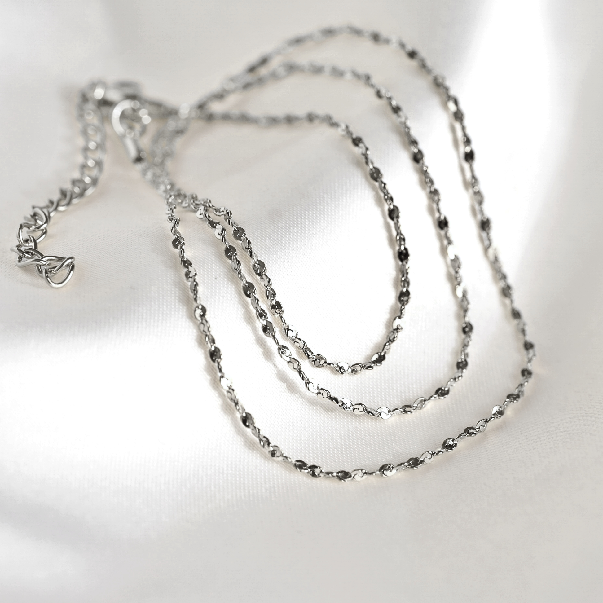 A silver-toned chain necklace with a shimmering rope link design, coiled on a white satin fabric background.