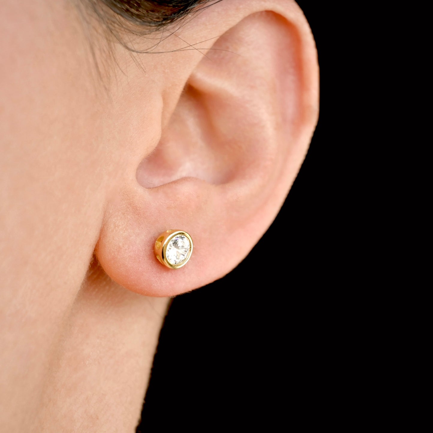 Gold solitaire stud earring with a clear gemstone worn by a person on a black background
