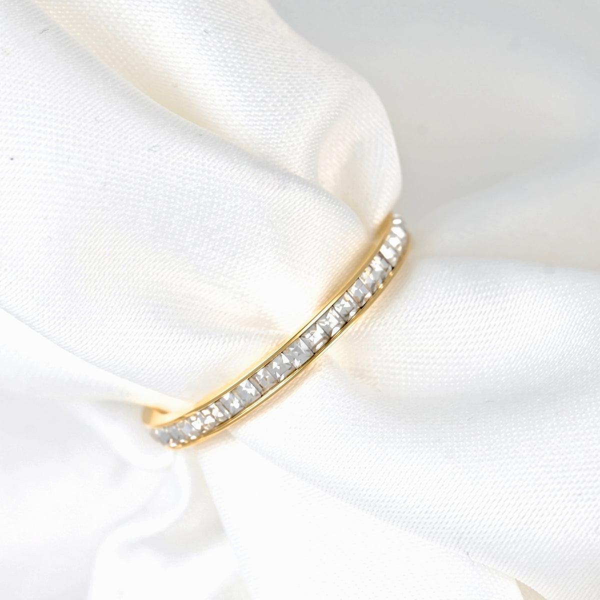 A gold eternity ring set with clear pavé gemstones, resting on a soft, draped white satin fabric.