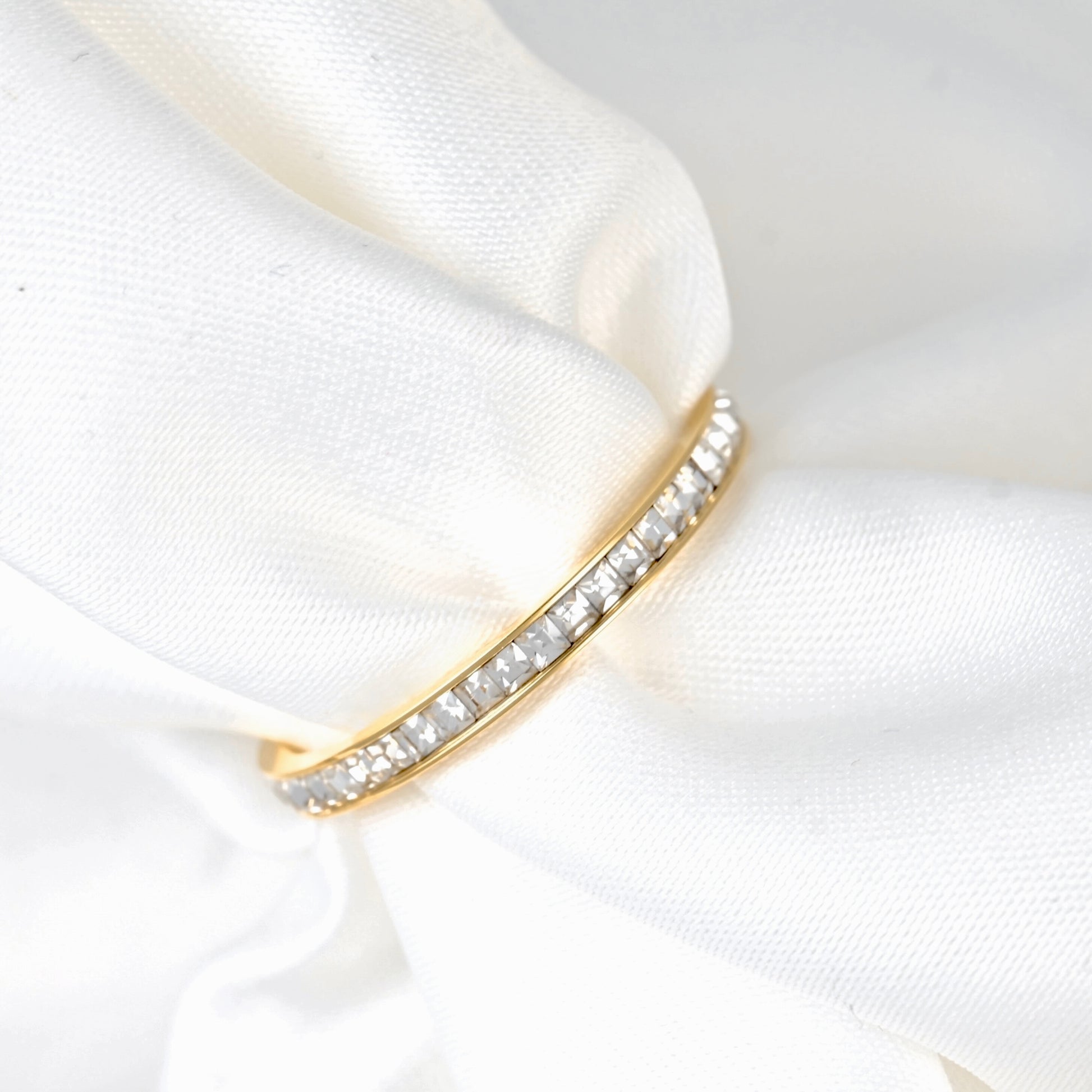 A gold eternity ring set with clear pavé gemstones, resting on a soft, draped white satin fabric.