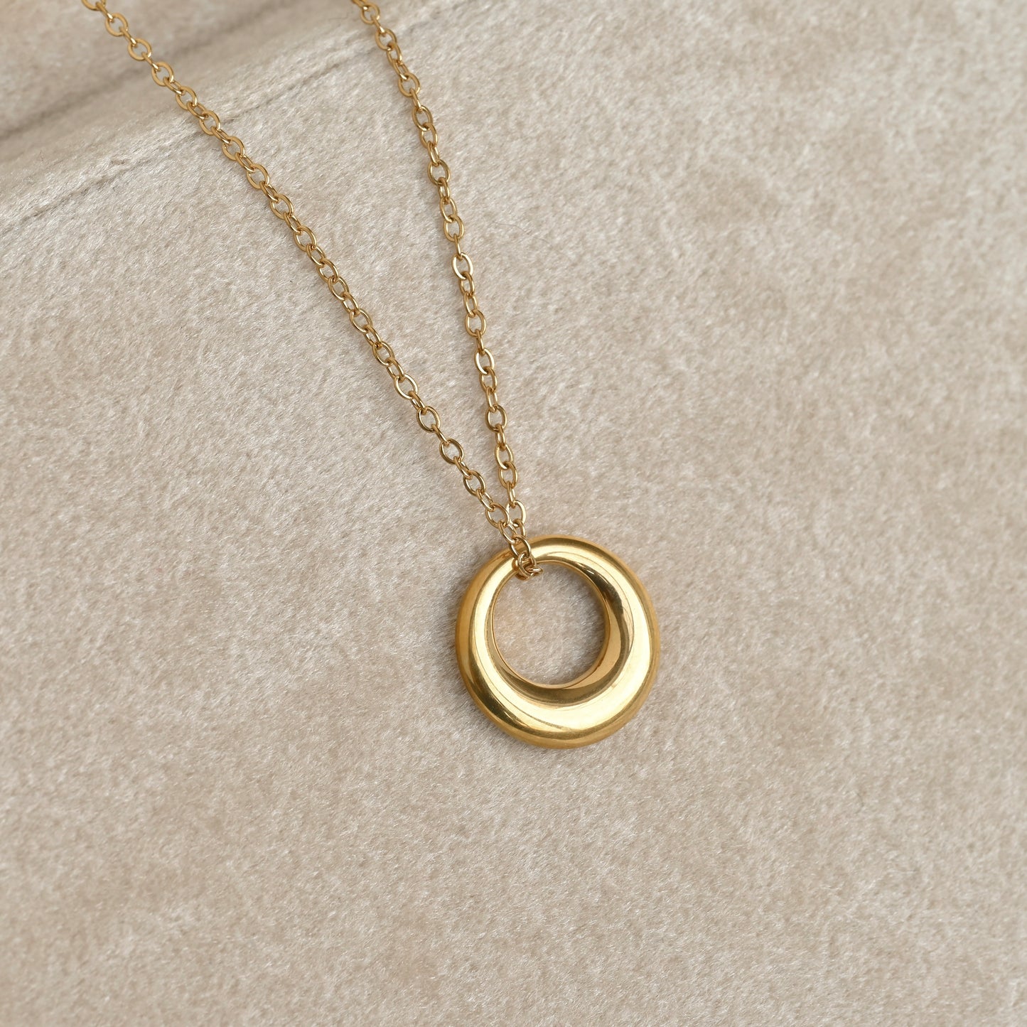 A gold chain necklace with a thick ring pendant, displayed on a textured beige fabric background.