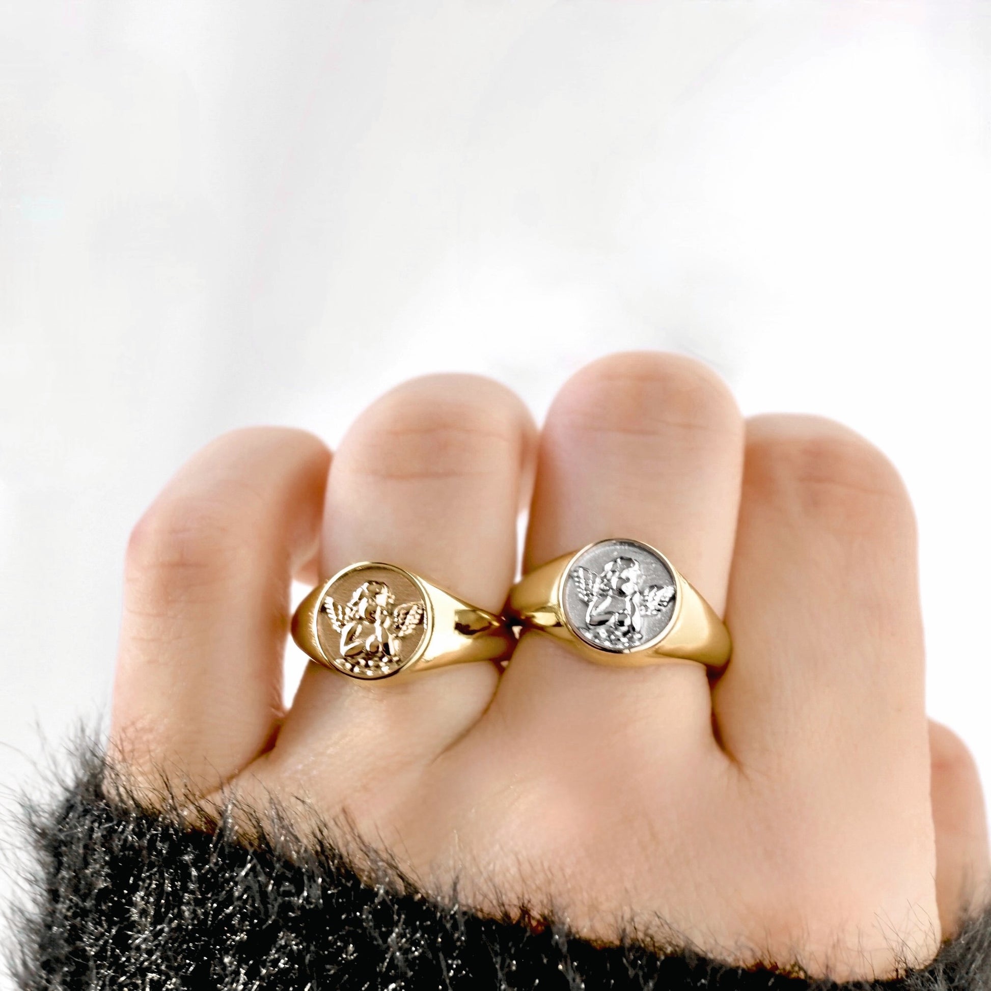 Two signet rings with round engraved faces worn on a hand. One ring is yellow gold and the other is two-tone, both featuring a raised angel emblem on a smooth band.