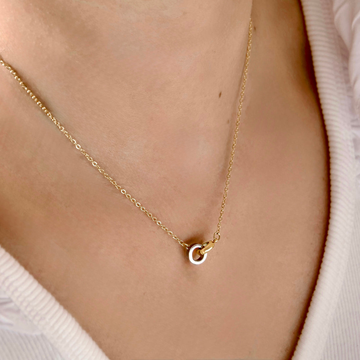 A delicate gold chain necklace with a small interlocking double-ring pendant, worn on a model's neck over a white ribbed top.