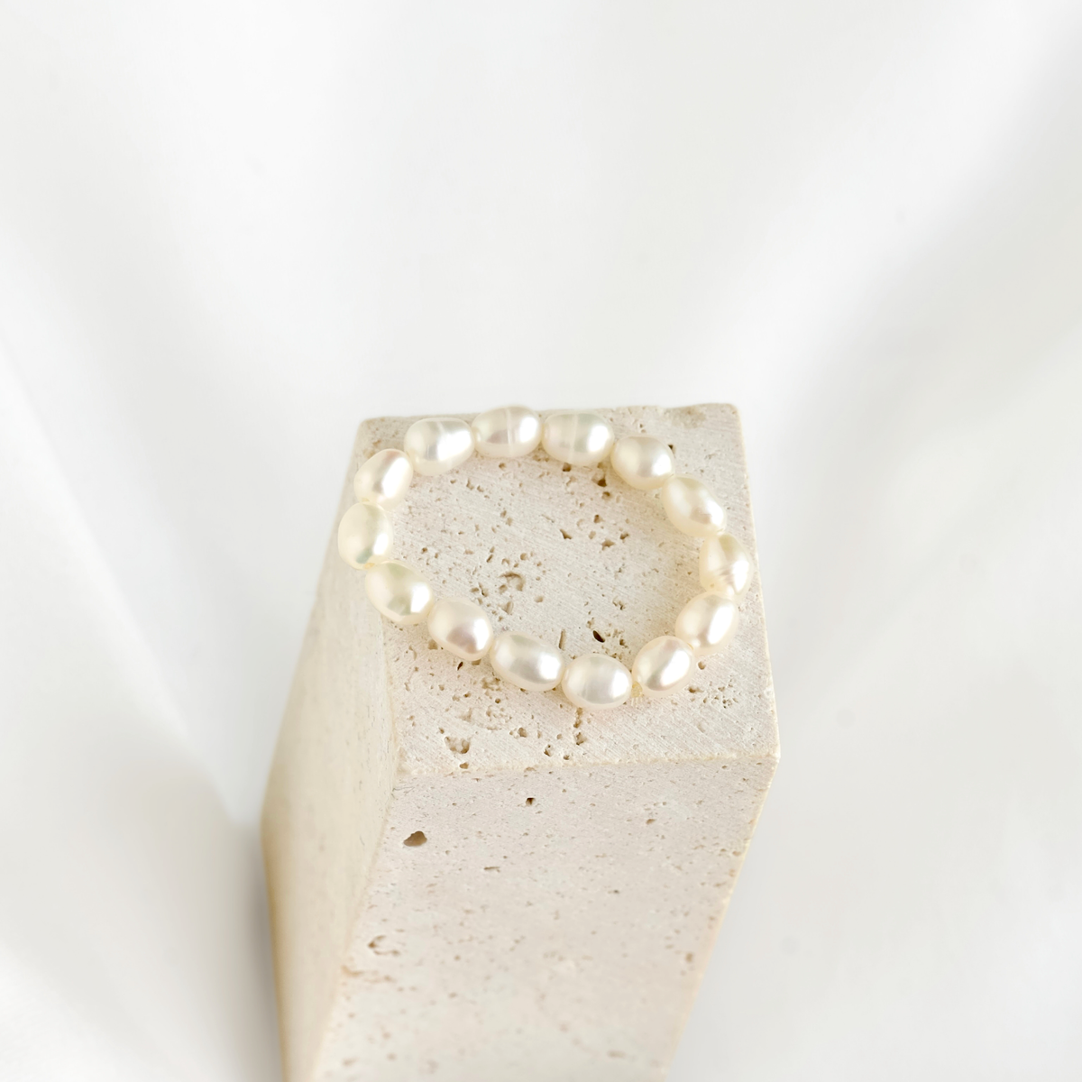 A beaded bracelet made of irregular white pearls, displayed on a textured beige stone block against a soft white background.