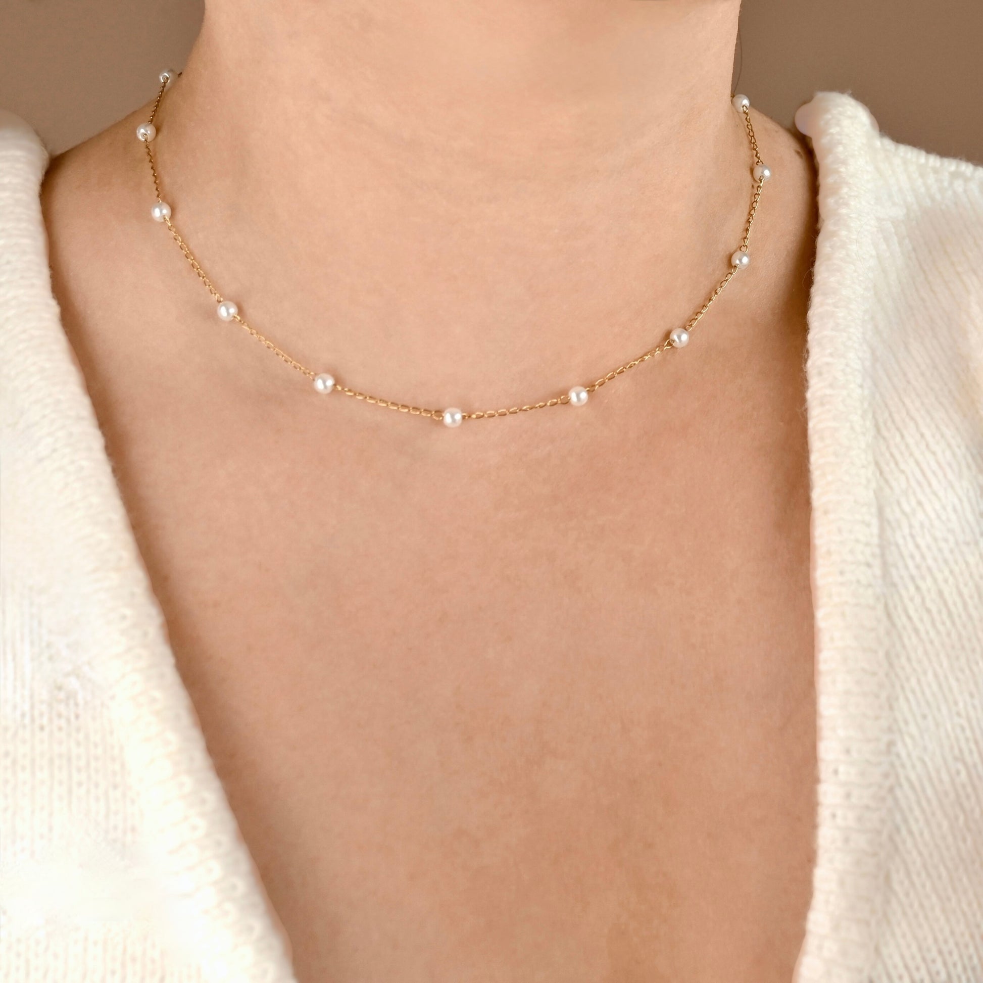 A delicate gold chain necklace with evenly spaced small white pearls, shown worn on a model's neck over a cream knit top.