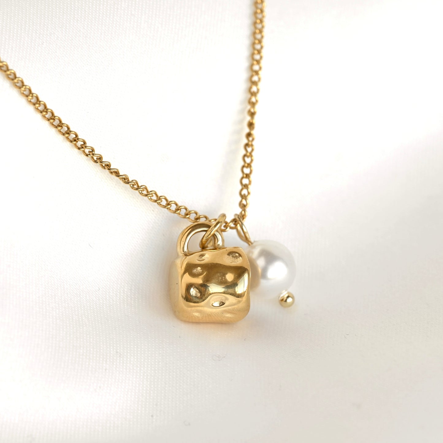 A gold chain necklace featuring a textured gold mug pendant with raised dot details and a small white pearl charm, displayed on a white fabric background.