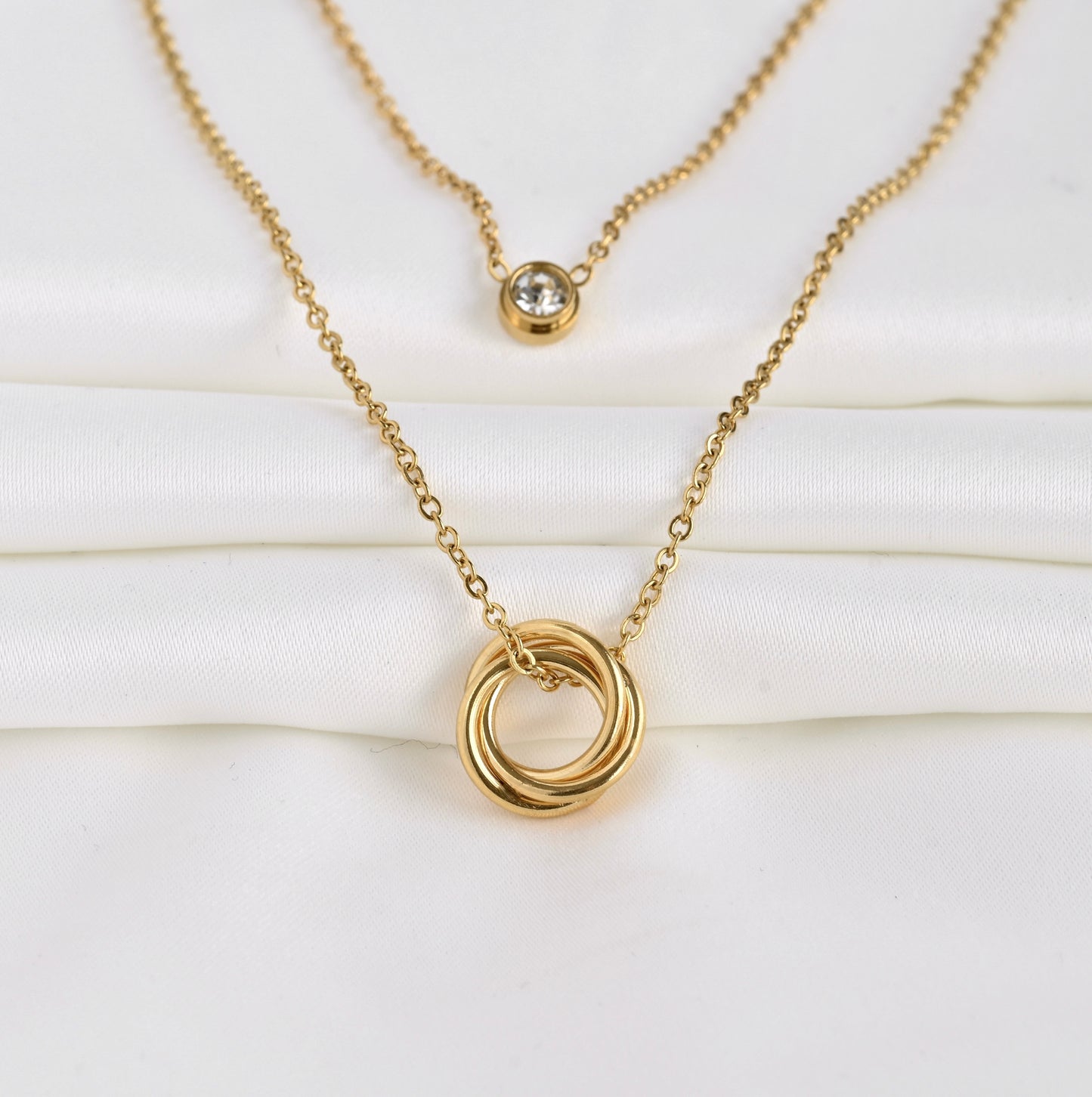 Wide angled front view of two layered gold necklace  set with interlocking rings and a bezel charm on a white background