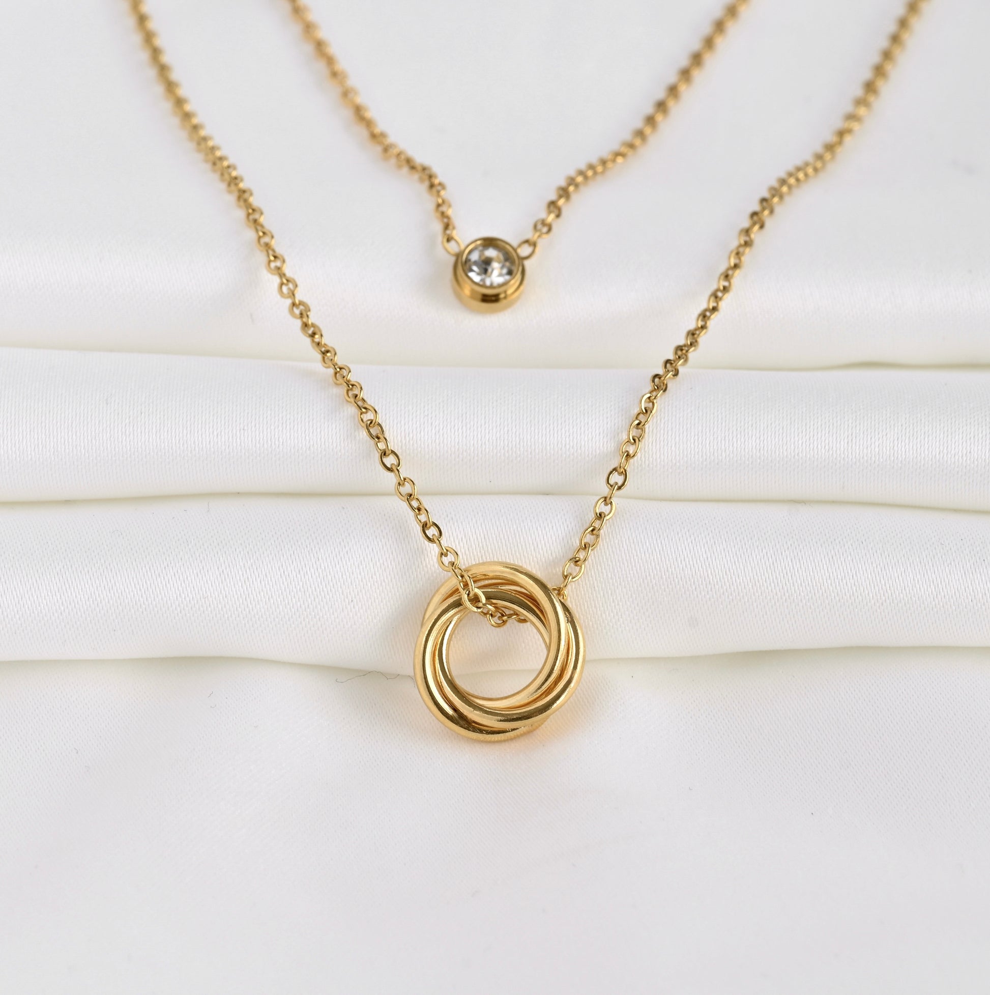 Wide angled front view of two layered gold necklace  set with interlocking rings and a bezel charm on a white background