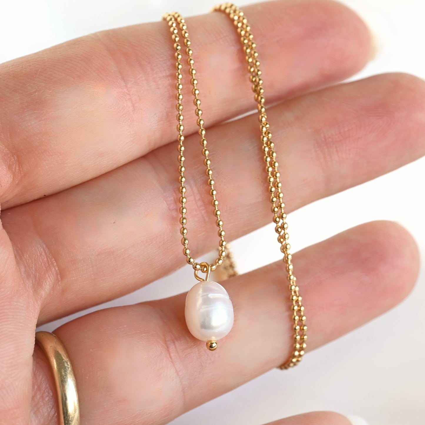 A delicate gold beaded chain necklace with a single irregular white baroque pearl pendant, held in a person's hand.