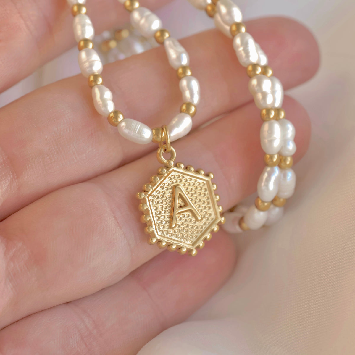 A close-up of a hand holding a beaded necklace with white baroque pearls, small gold spacer beads, and a textured gold hexagonal pendant engraved with the letter A.