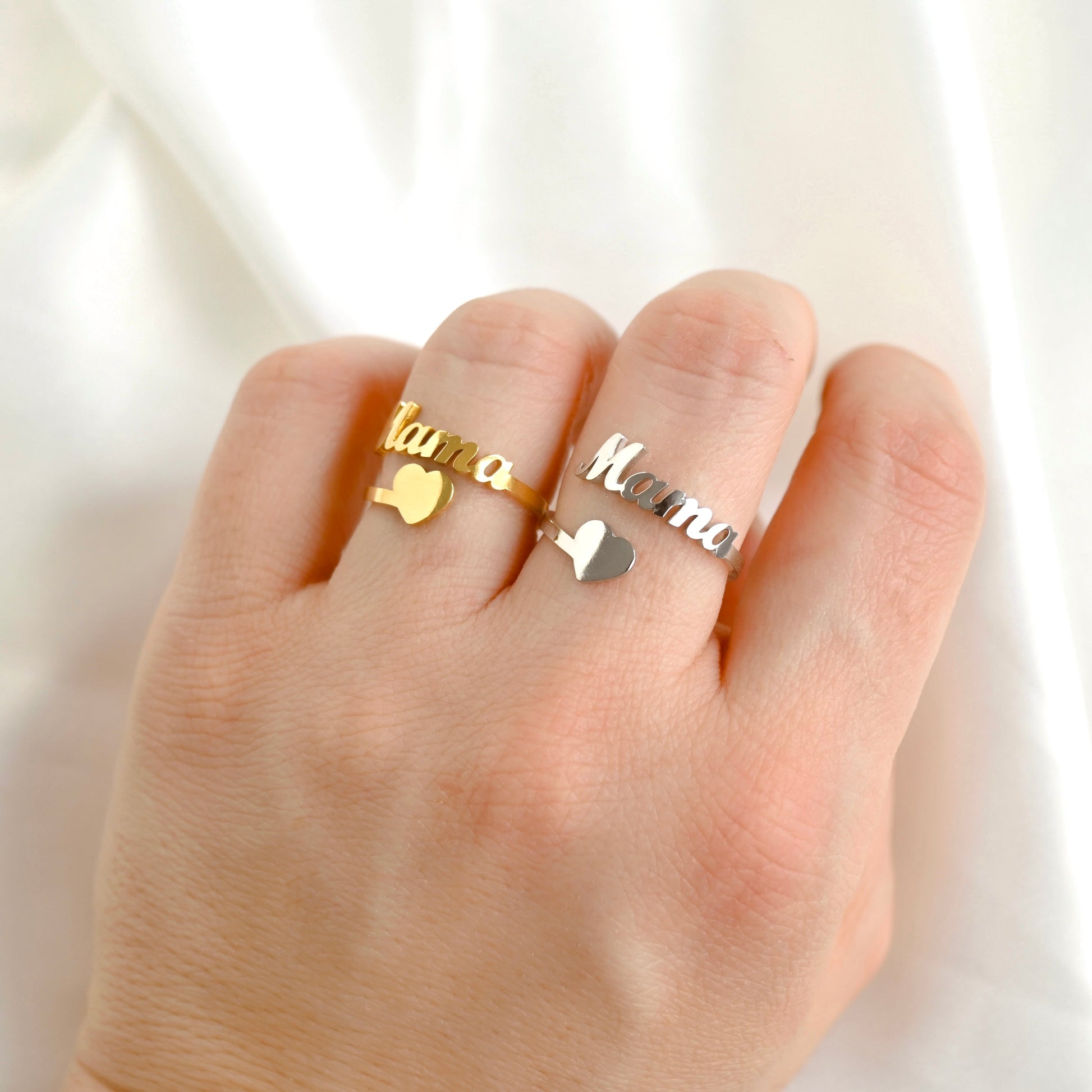 A close-up of a hand wearing two slim metal rings with heart charms and cursive 'Mama' nameplate, one in gold and one in silver, against a soft white fabric background.