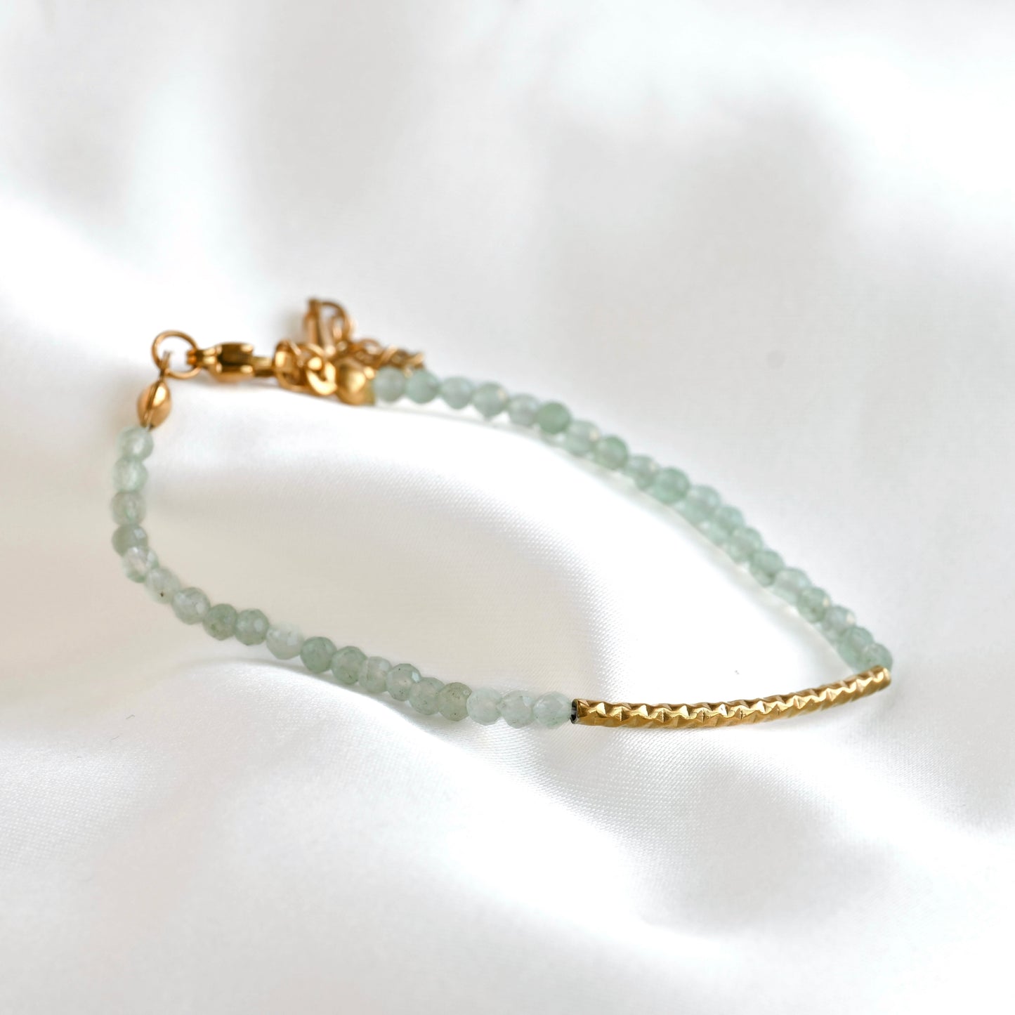 A dainty green beaded bracelet featuring pale green faceted beads and gold-toned metal accents, including a textured bar and clasp, displayed on a soft white fabric background.