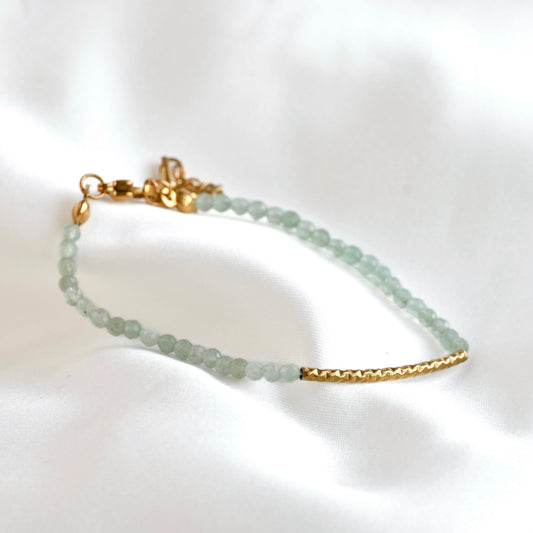 A dainty green beaded bracelet featuring pale green faceted beads and gold-toned metal accents, including a textured bar and clasp, displayed on a soft white fabric background.