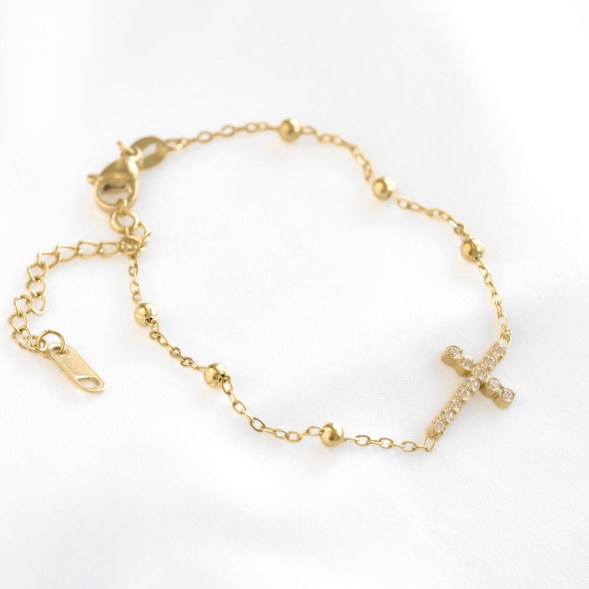 A delicate gold chain bracelet featuring a small cross charm paved with clear stones, accented with small gold beads, displayed on a clean white background.
