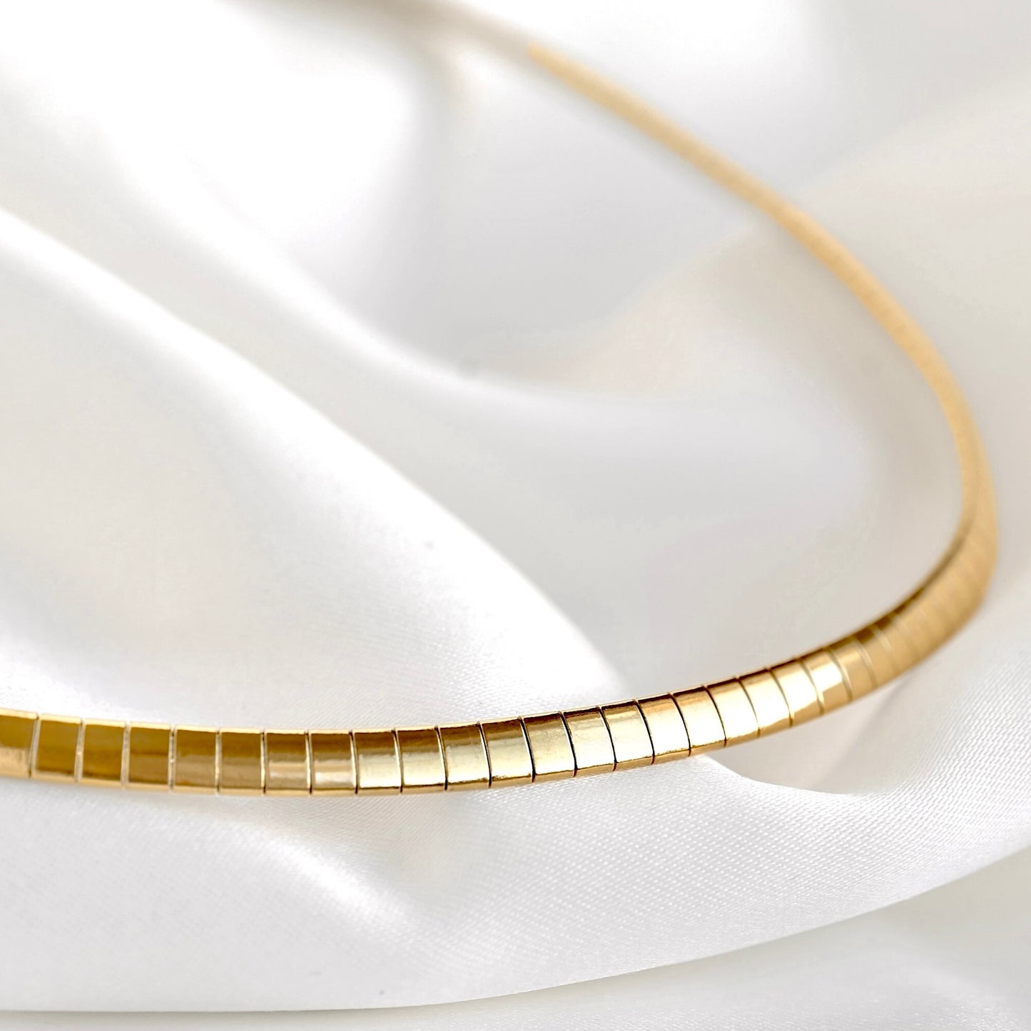 A close-up of a gold omega chain necklace draped over soft white satin fabric.