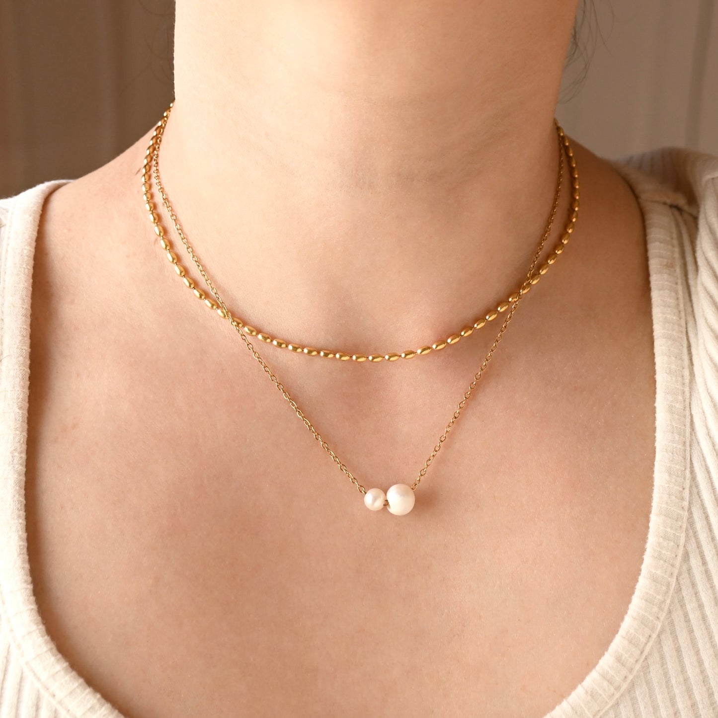 A close-up of a person's neck and chest wearing a double-layered gold necklace. The top layer features an puffy oval link chain, and the bottom layer is a delicate gold chain with one big and one small white freshwater pearls at the center.