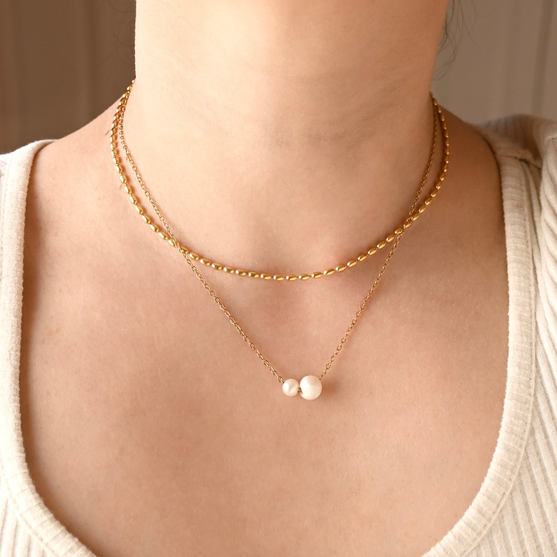 A close-up of a person's neck and chest wearing a double-layered gold necklace. The top layer features an puffy oval link chain, and the bottom layer is a delicate gold chain with one big and one small white freshwater pearls at the center.