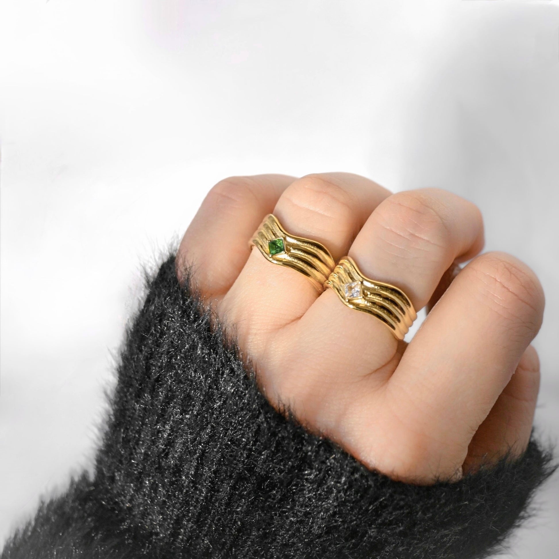 A close-up of a hand wearing two gold wavy band rings, one with a small green gemstone and the other with a small clear gemstone, set against a soft white background with a dark fuzzy sleeve visible.