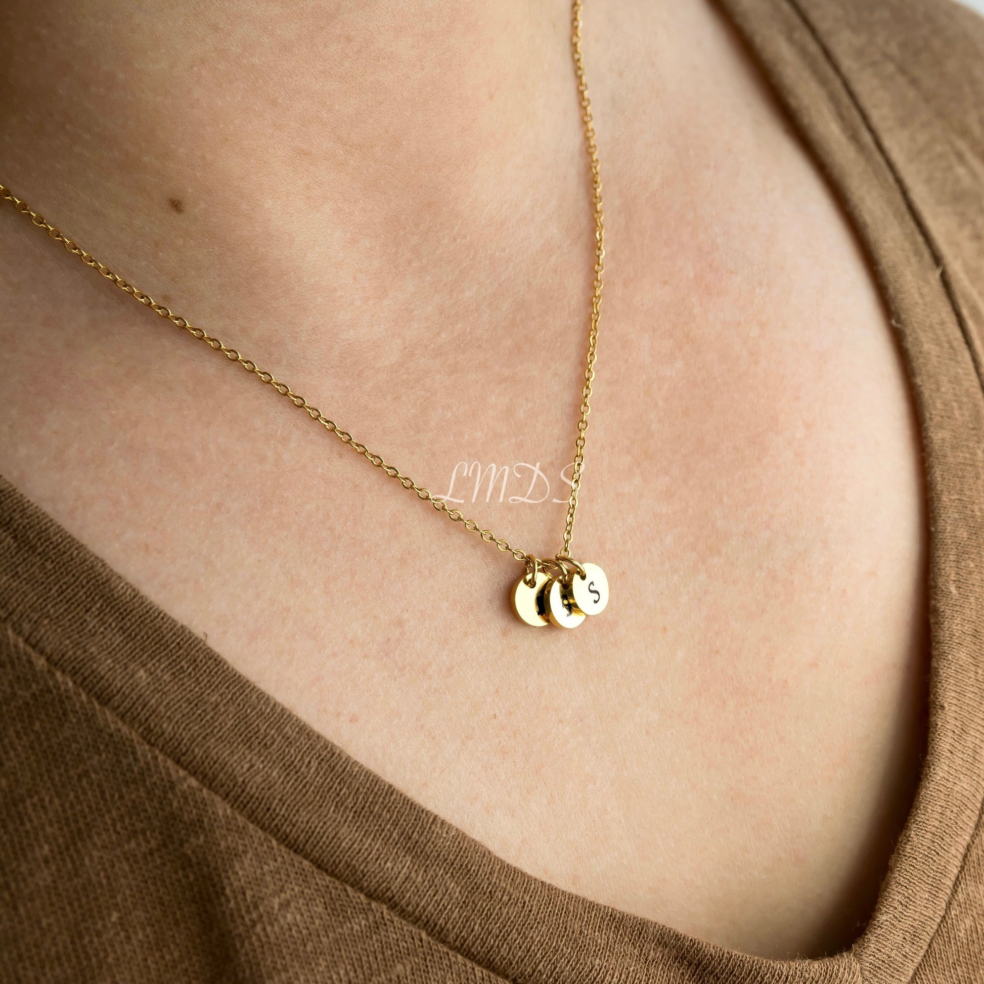Gold necklace with three round initial charms on a person wearing a brown top.