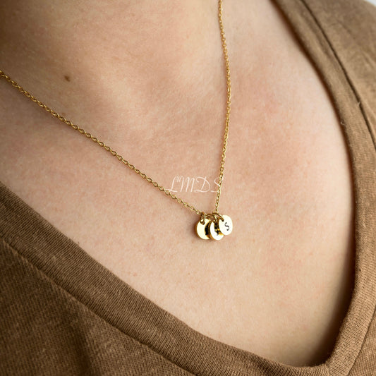 Gold necklace with three round initial charms on a person wearing a brown top.