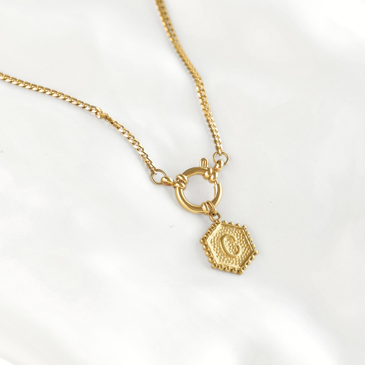 A gold chain necklace featuring a toggle clasp and a small hexagonal pendant with an embossed letter 'G', displayed on a soft white fabric background.