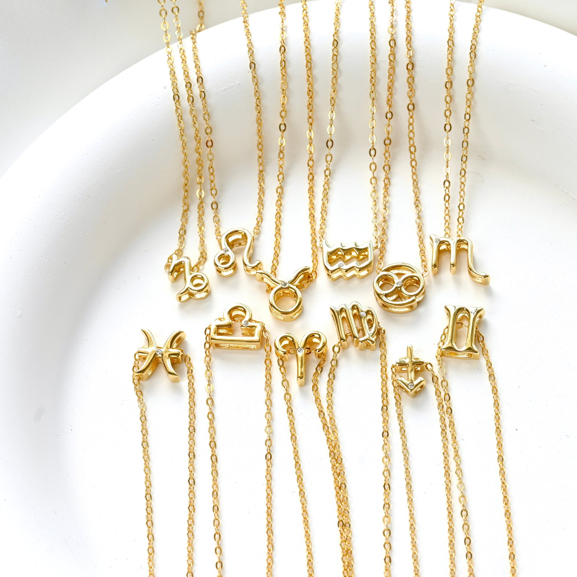 Twelve gold necklaces with zodiac symbol pendants in a white dish.