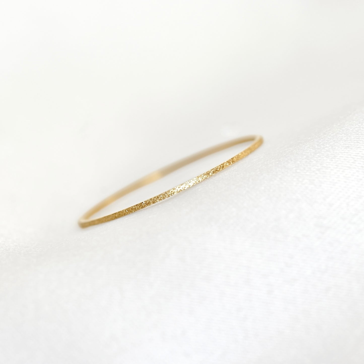A delicate, thin gold ring with a textured, glittery finish, resting on a soft white fabric surface.
