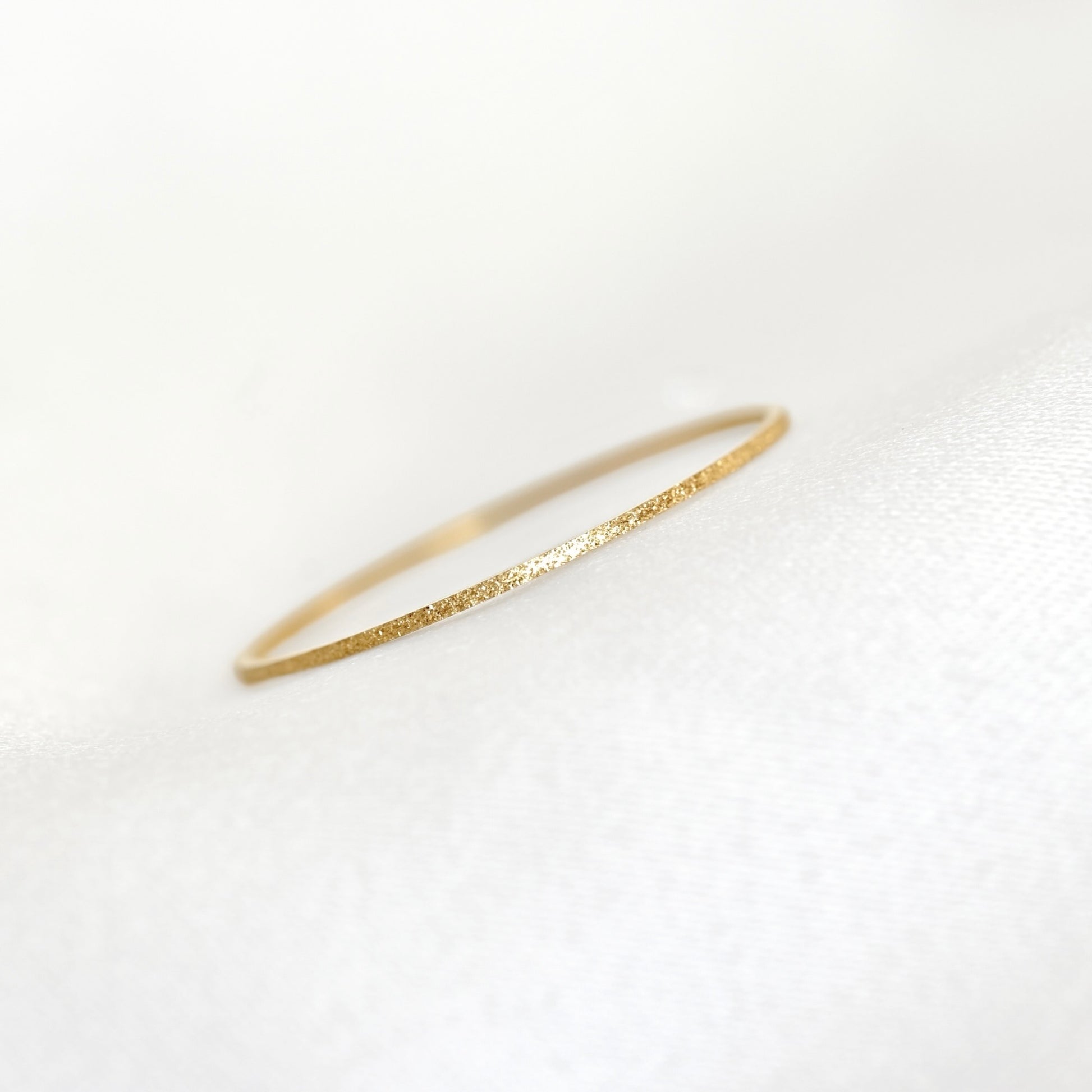 A delicate, thin gold ring with a textured, glittery finish, resting on a soft white fabric surface.