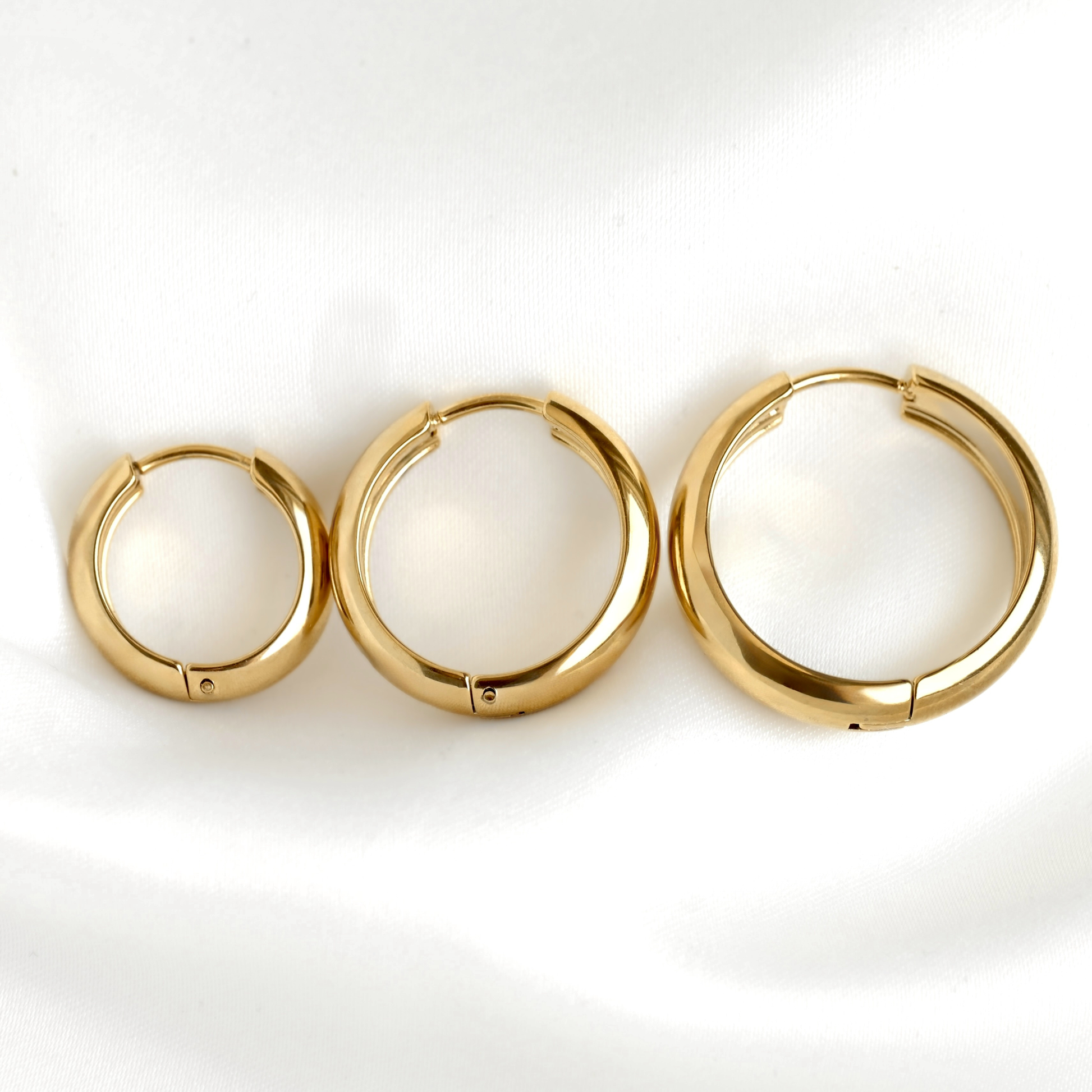 Three gold hoop earrings in different sizes arranged horizontally on a white fabric background.