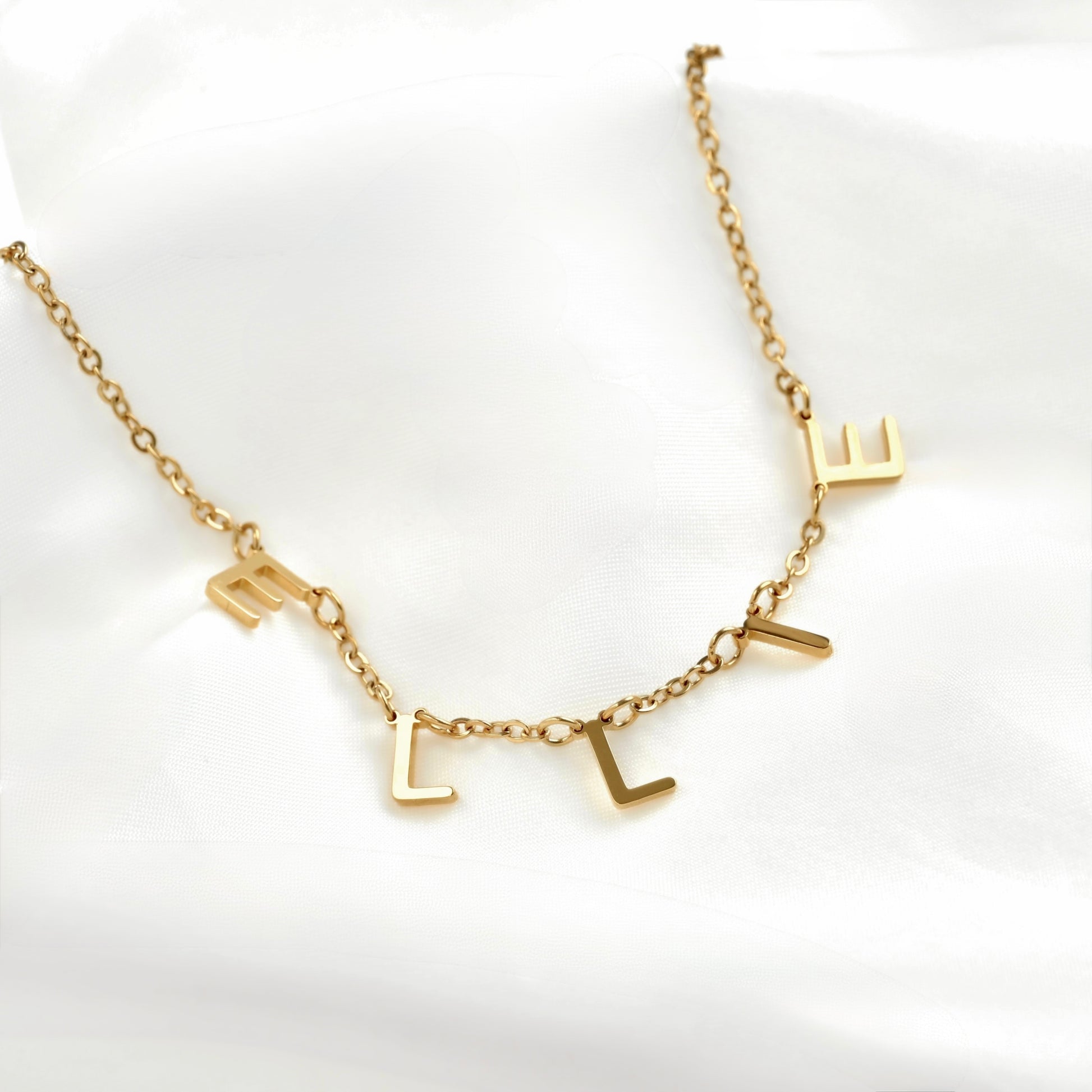A gold initial necklace spelling 'E L L I E' in a delicate cable chain, displayed on a soft white fabric background.