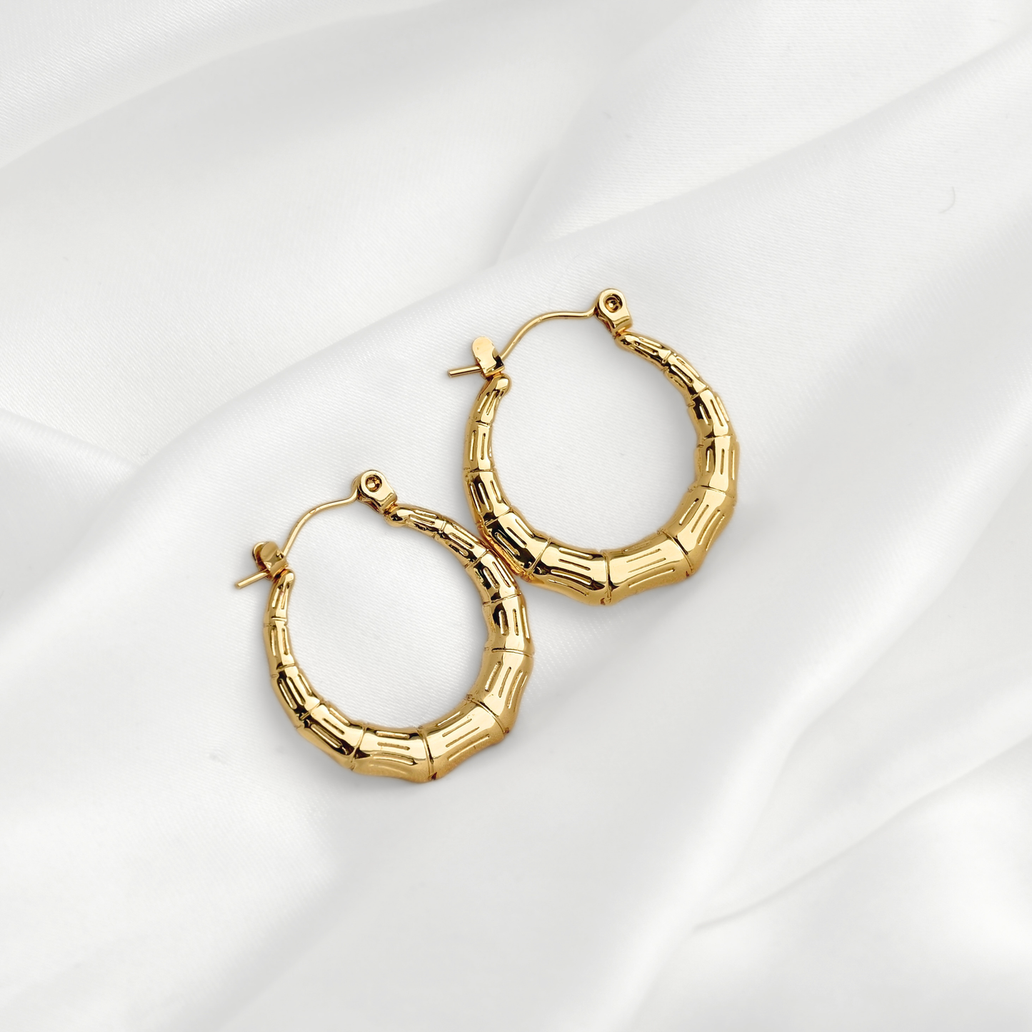 A pair of gold hoop earrings with a textured, segmented bamboo-like design, displayed on a soft white fabric background.