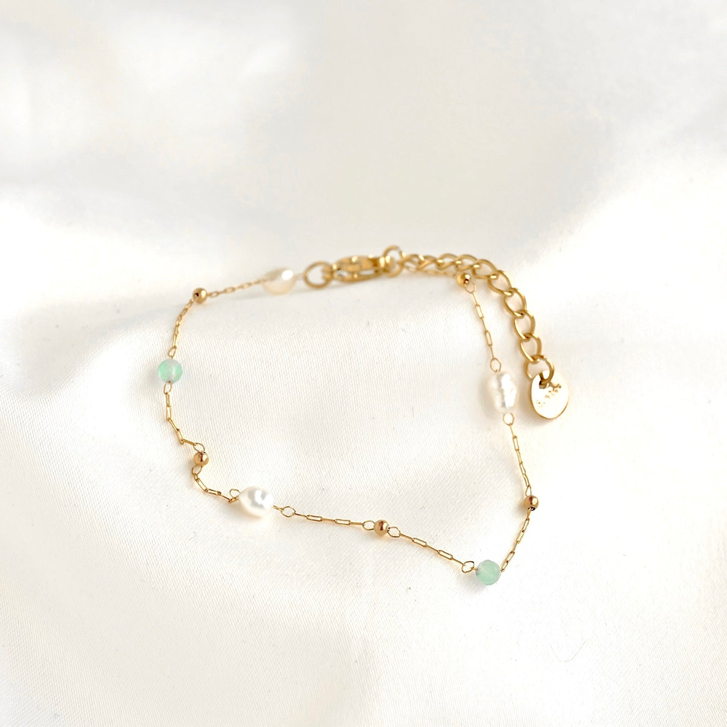 A delicate gold chain bracelet with small white pearls and light green beads, displayed on a soft white fabric background.
