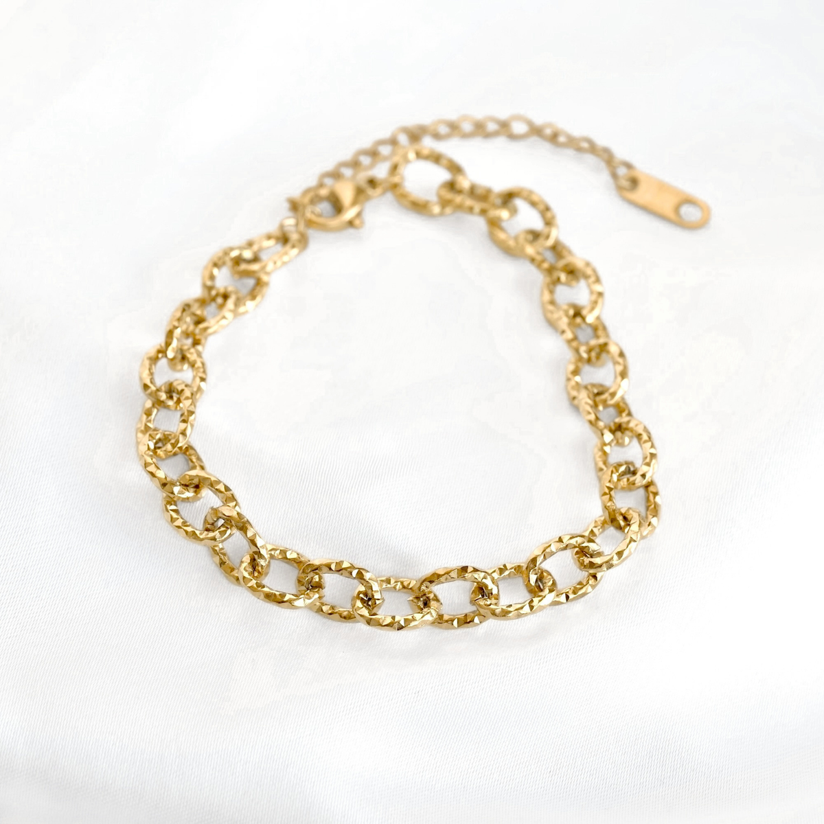 A gold-toned chain bracelet with textured oval links and an adjustable lobster clasp, displayed on a white fabric background.