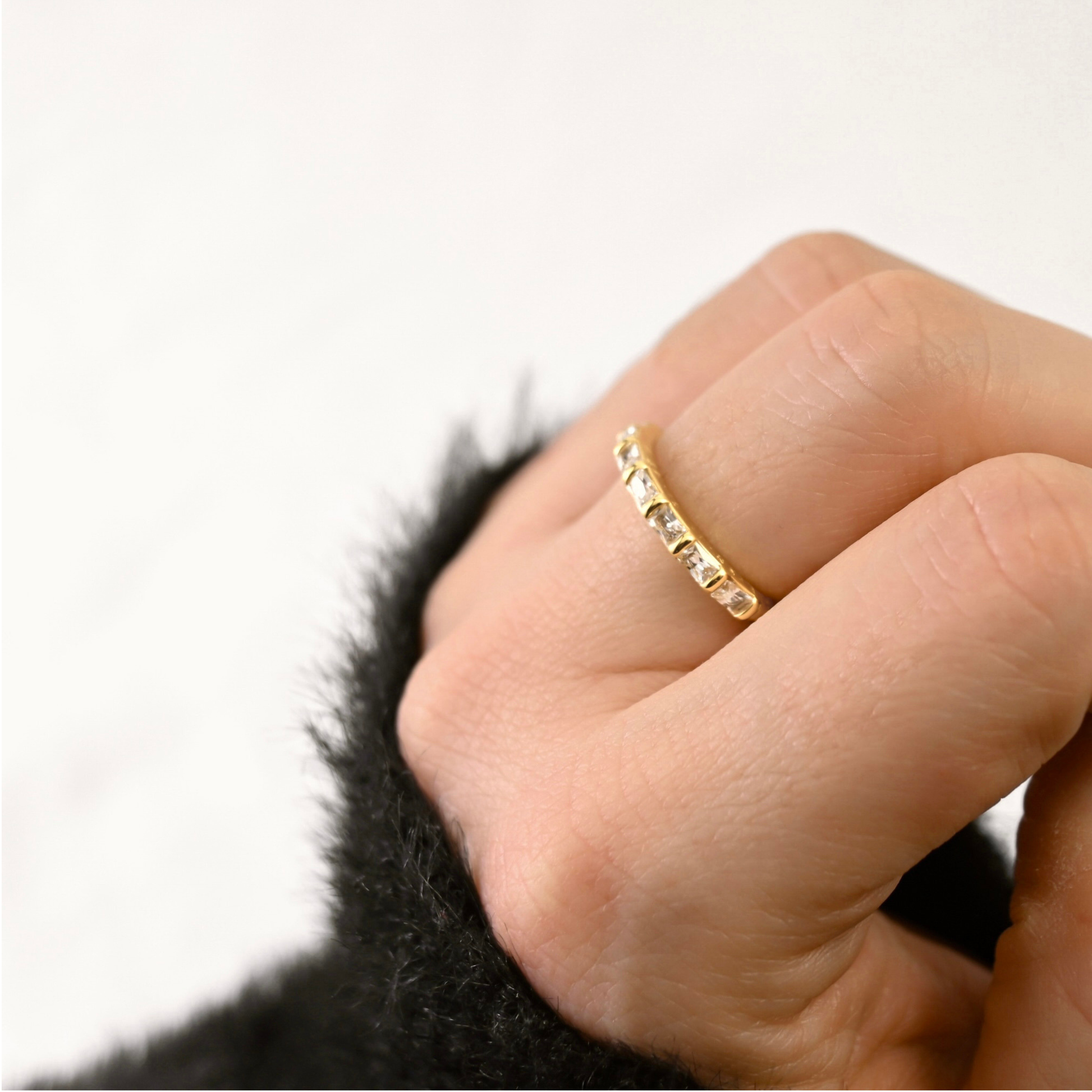 A close-up of a hand wearing a delicate gold band ring with small clear stones, set against a soft white background with a dark fuzzy sleeve visible at the bottom.