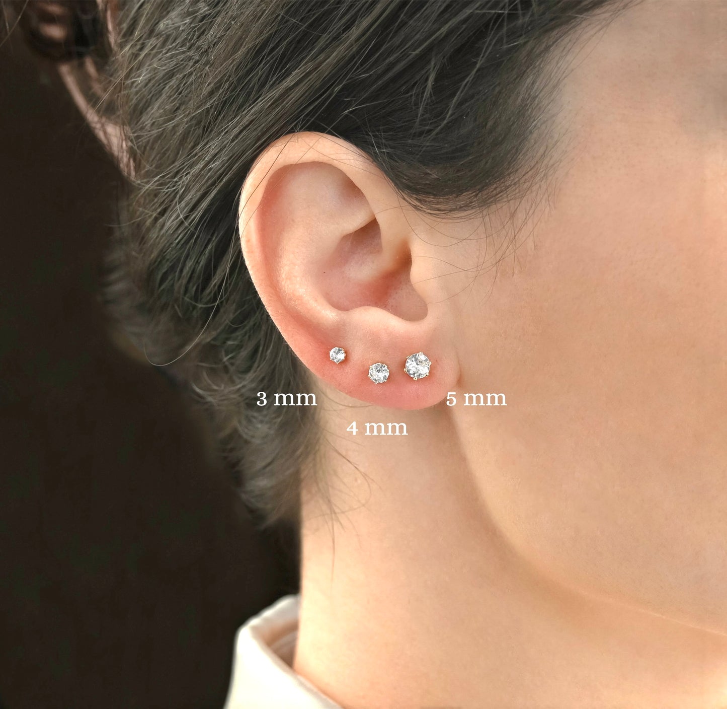 Close-up of an ear wearing three gold CZ stud earrings with size labels '3mm', '4mm and '5mm'