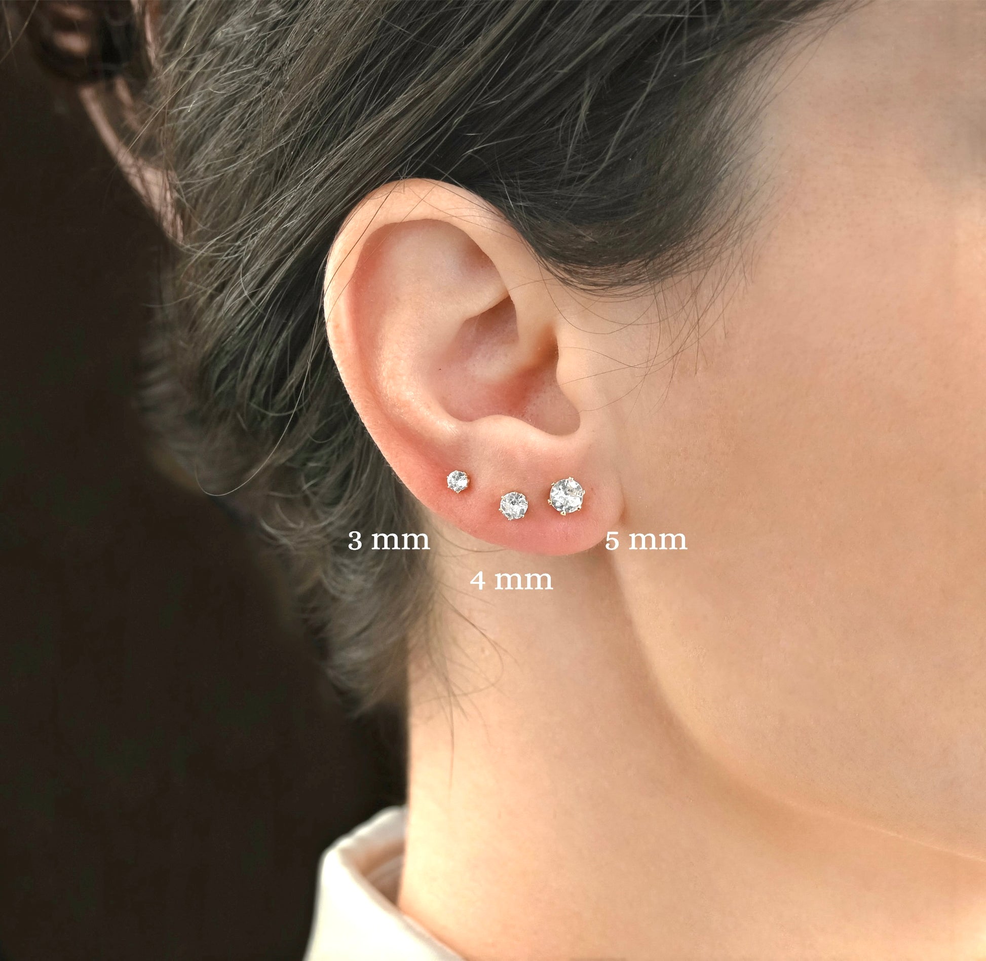 Close-up of an ear wearing three gold CZ stud earrings with size labels '3mm', '4mm and '5mm'