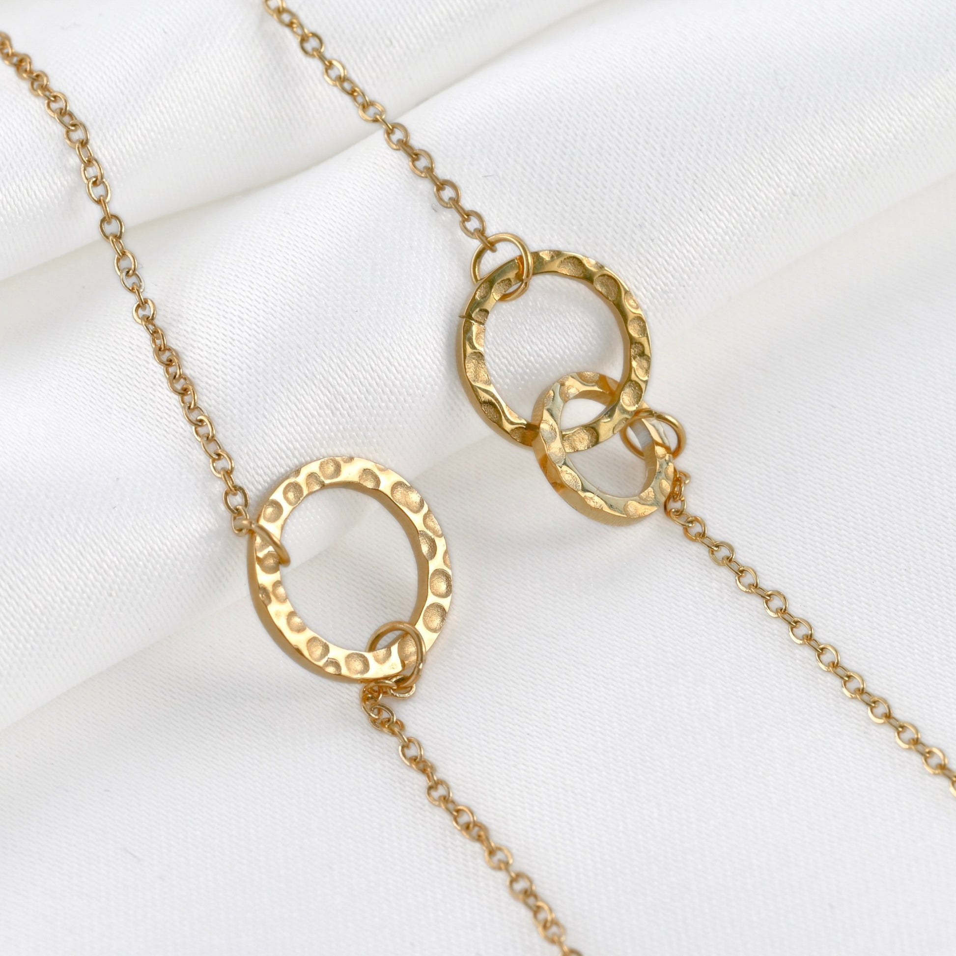 Two gold bracelets with a single circle and interlocking circles on a delicate cable chain on white satin.