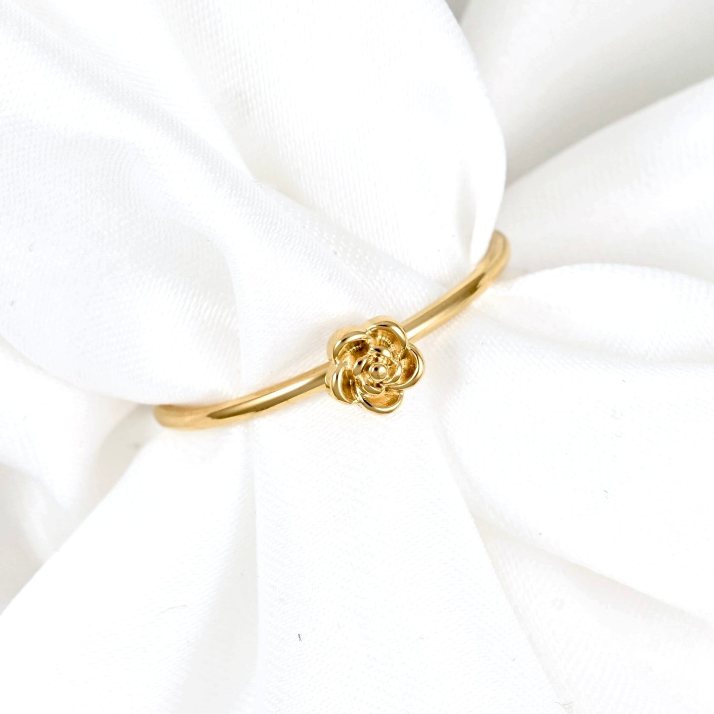 A delicate gold band ring featuring a small rose-shaped charm in the center, displayed on soft white fabric.