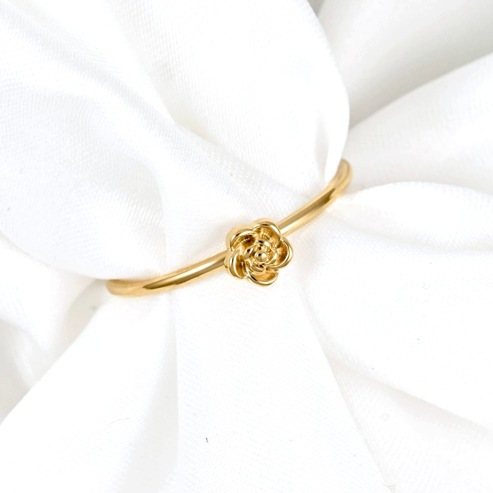 A delicate gold band ring featuring a small rose-shaped charm in the center, displayed on soft white fabric.